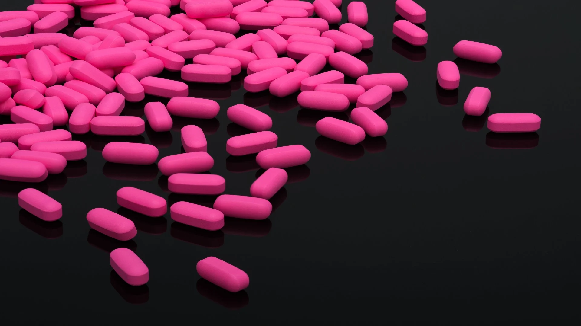 Pink pill overdose risk illustrated with scattered pink tablets on a dark reflective surface.