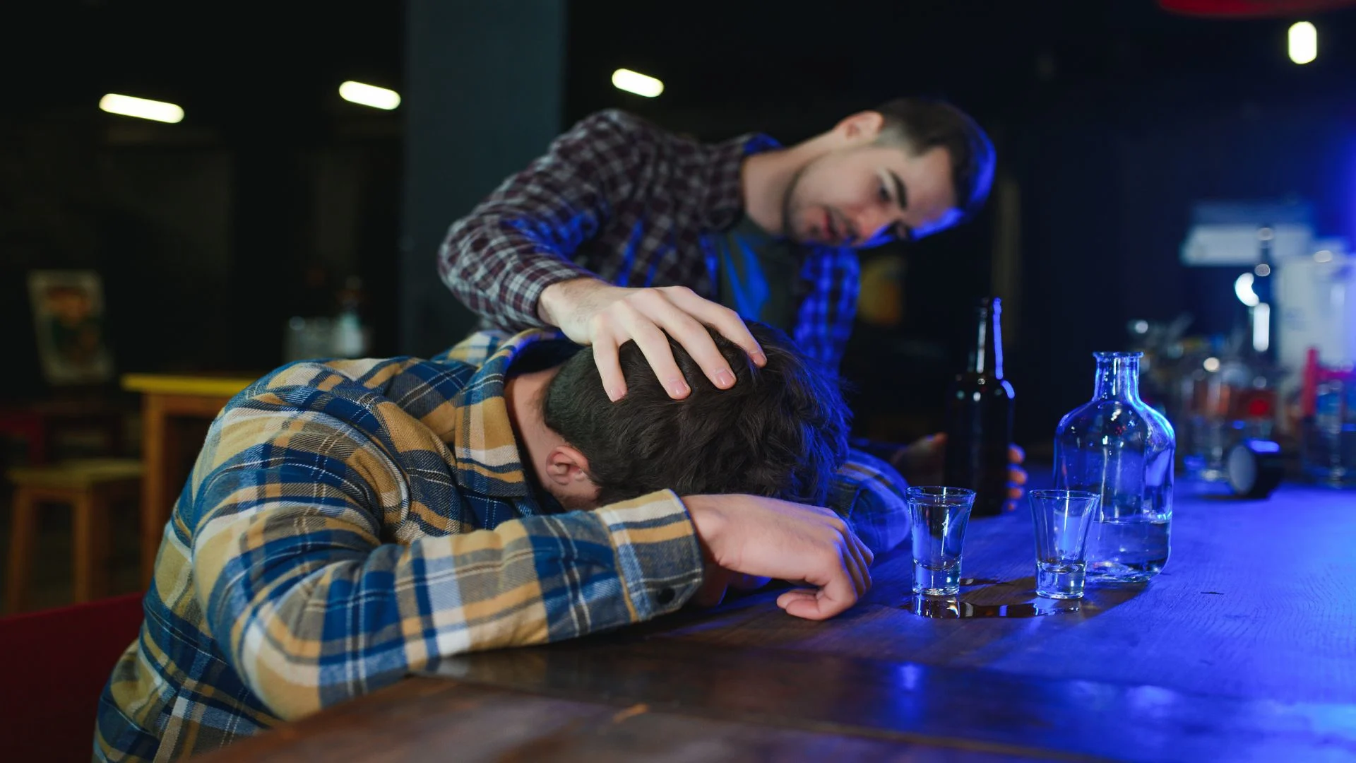 alcohol poisoning at a bar where a man is checking on his passed out friend on the bar counter in an interesting blue lighting scene.