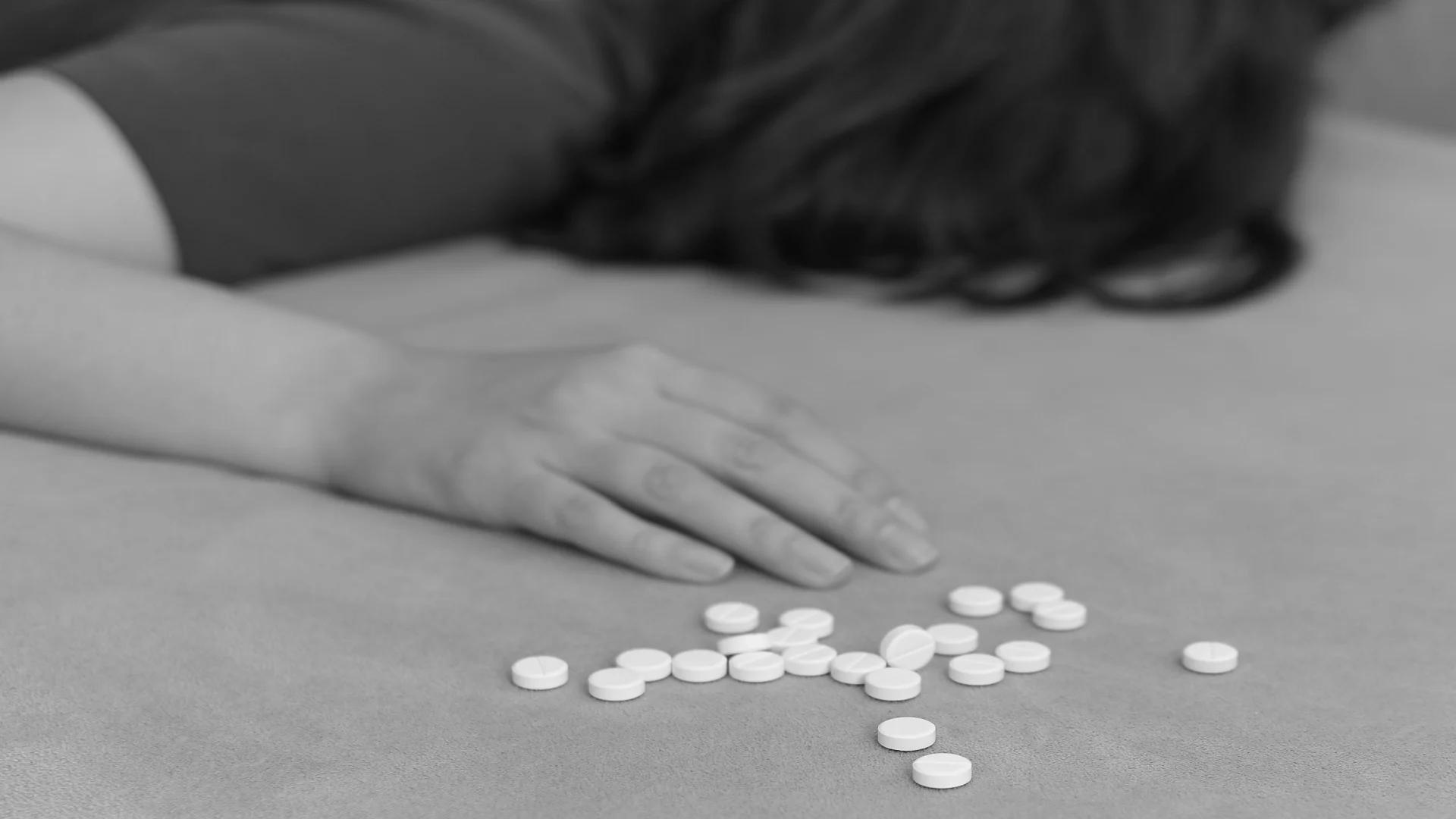 Scene depicting opioid overdose with a woman passed out near a scattered pile of white pills on a bed, in black and white.