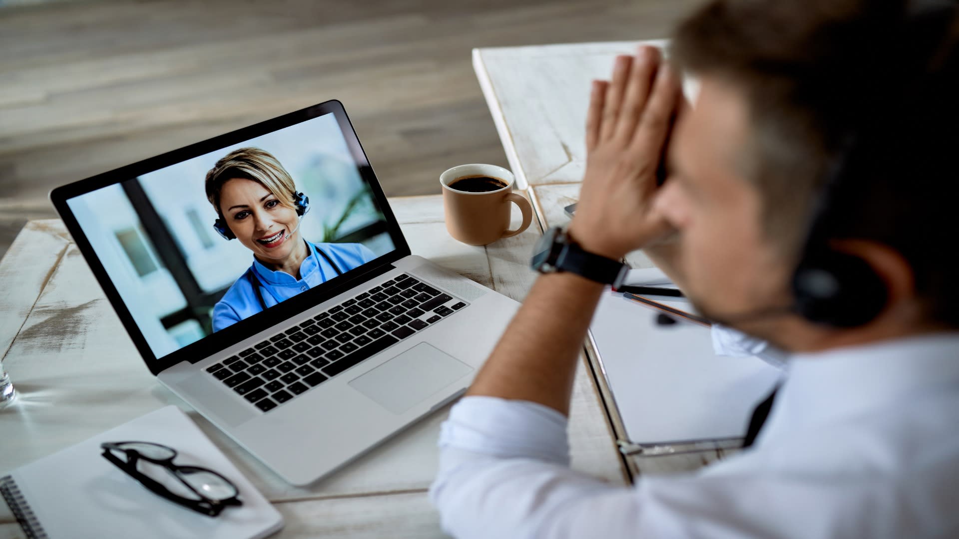 Stressed man attending a telehealth mental health video call with a smiling female therapist on a laptop screen.