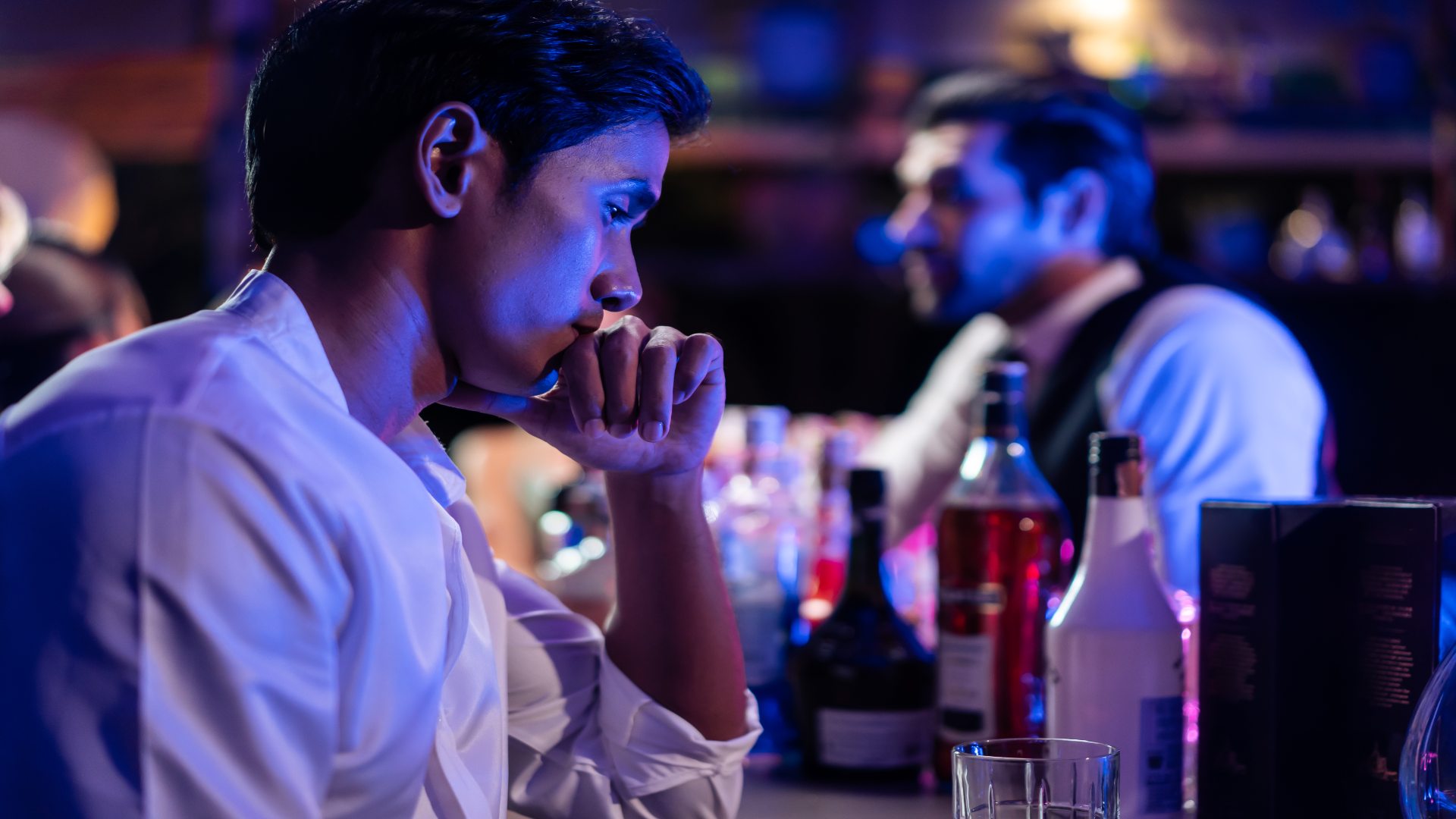 Alcohol intoxication symptoms depicted in a young man sitting pensively at a bar, surrounded by bottles and dim lighting, suggesting emotional and cognitive impairment.