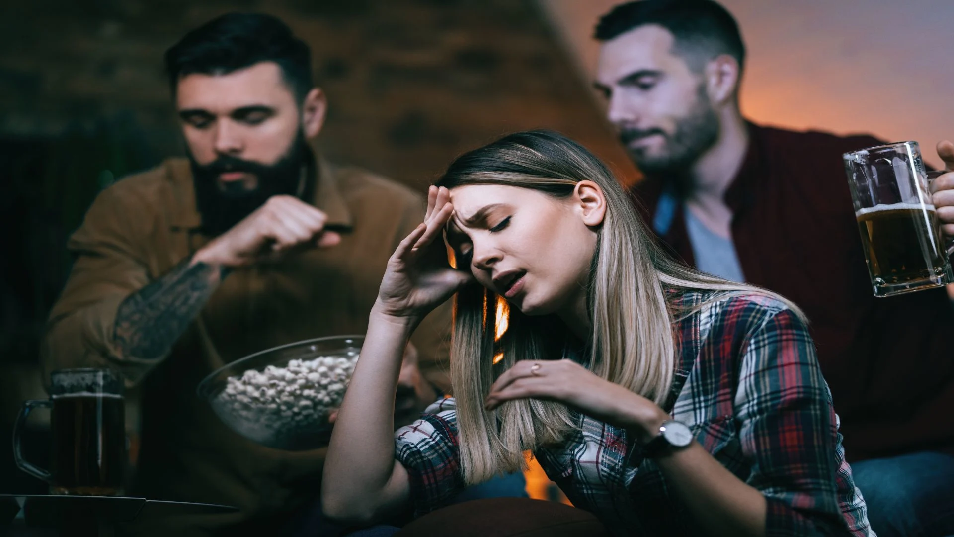 A woman holds her head in discomfort while surrounded by people drinking alcohol, symbolizing social stress and emotional strain—common meth withdrawal symptoms.