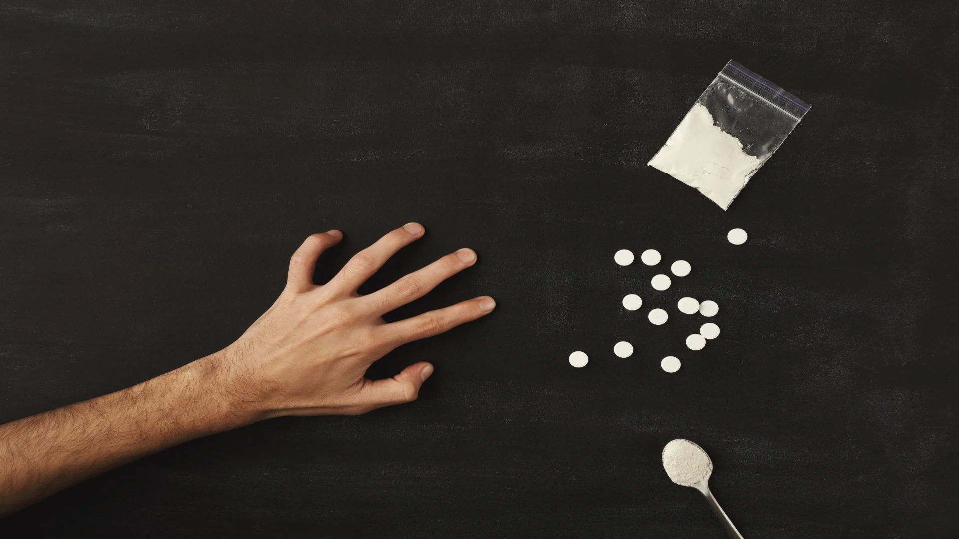 Outstretched hand reaching for pills and white powder on a black surface — symbolizing drug misuse and the risks of benzodiazepine abuse such as Valium.