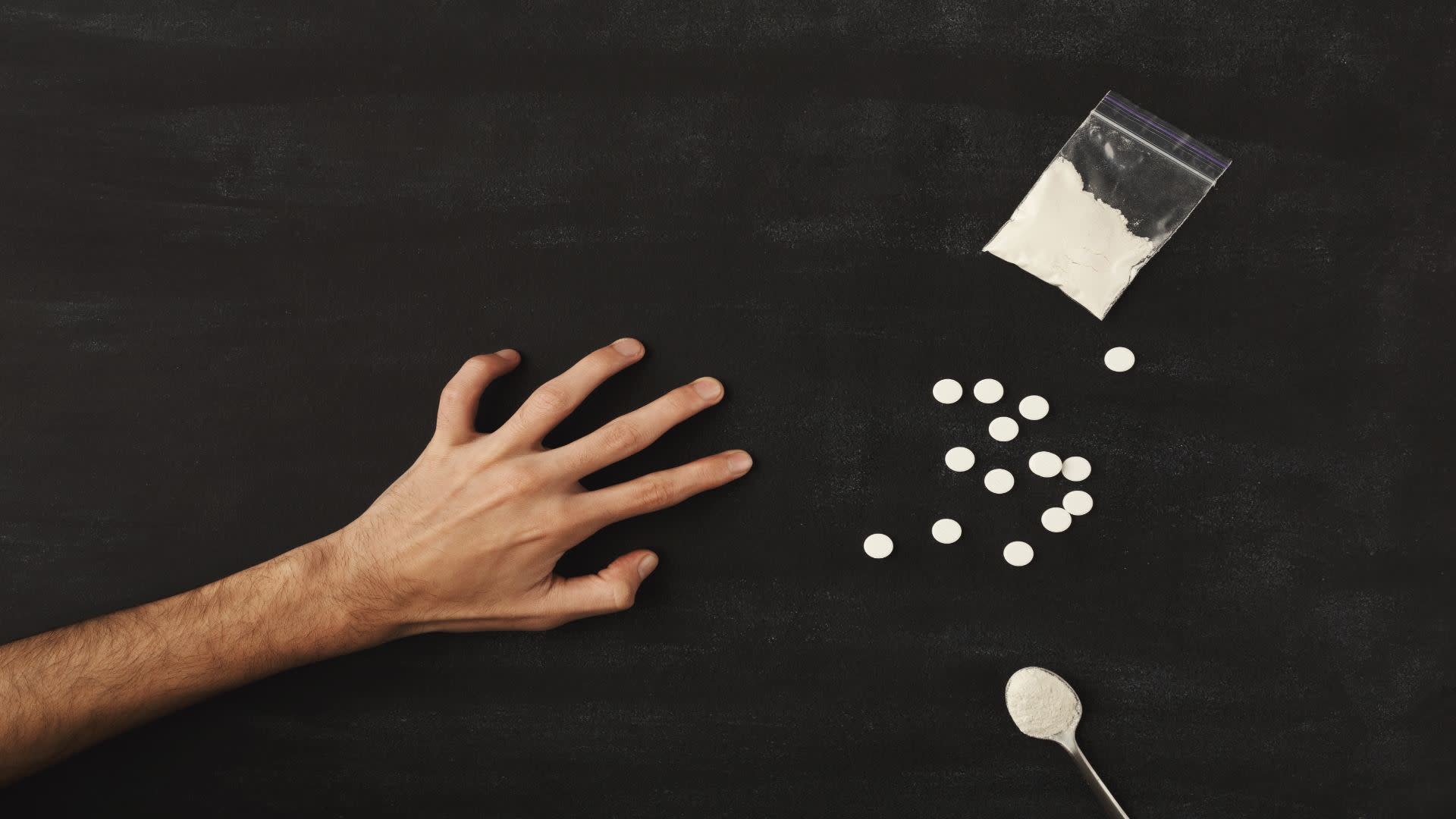 Outstretched hand reaching for pills and white powder on a black surface — symbolizing drug misuse and the risks of benzodiazepine abuse such as Valium.