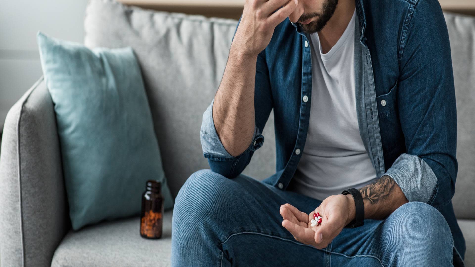 A distressed man sitting on a couch with a handful of pills, reflecting potential misuse of prescription drugs. A relevant visual for articles exploring what are benzodiazepines and their risks.
