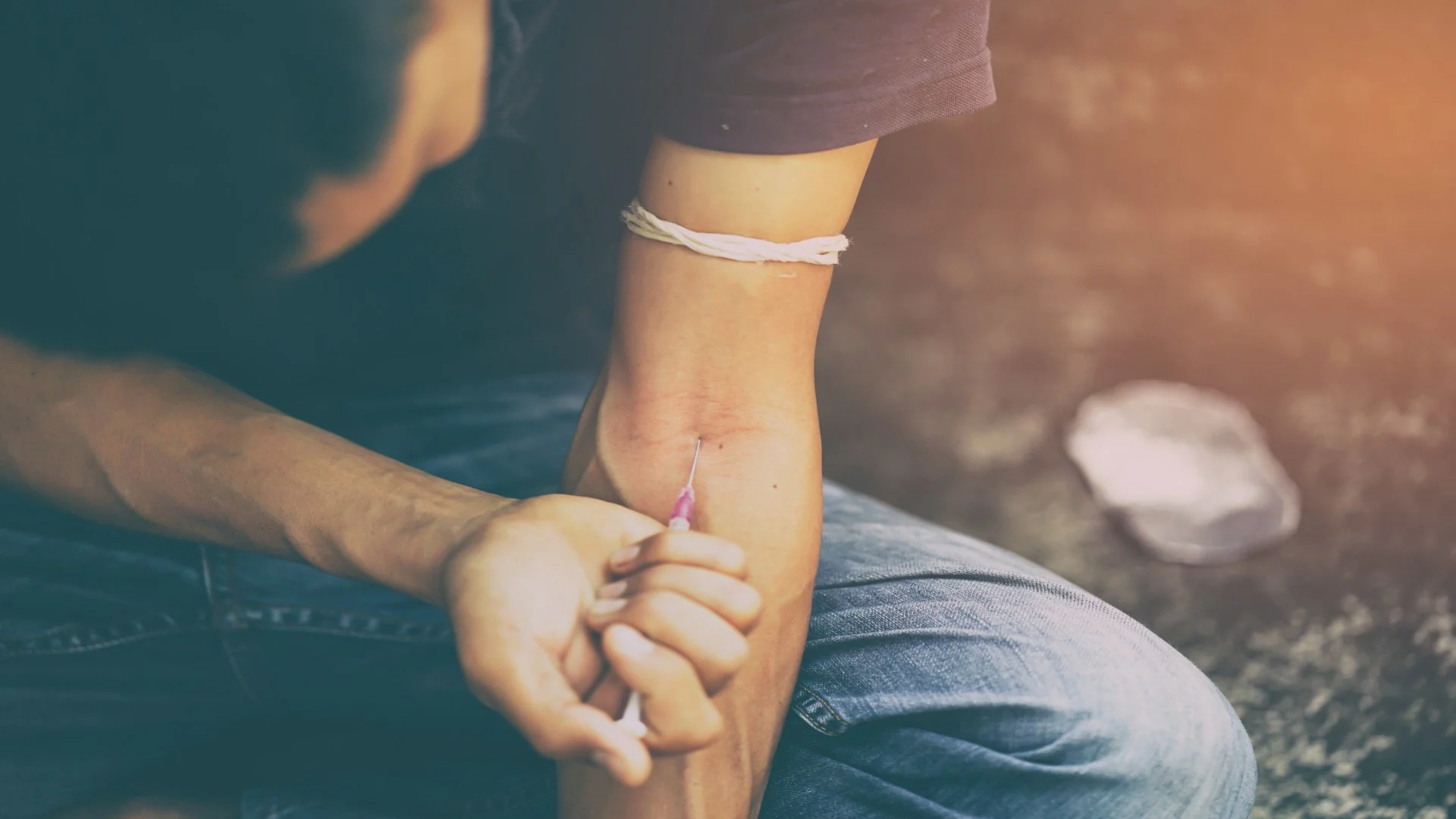 Close-up of a person injecting drugs into their arm while sitting on the ground, with a makeshift tourniquet tied around the bicep — illustrating the desperation and physical toll of addiction.