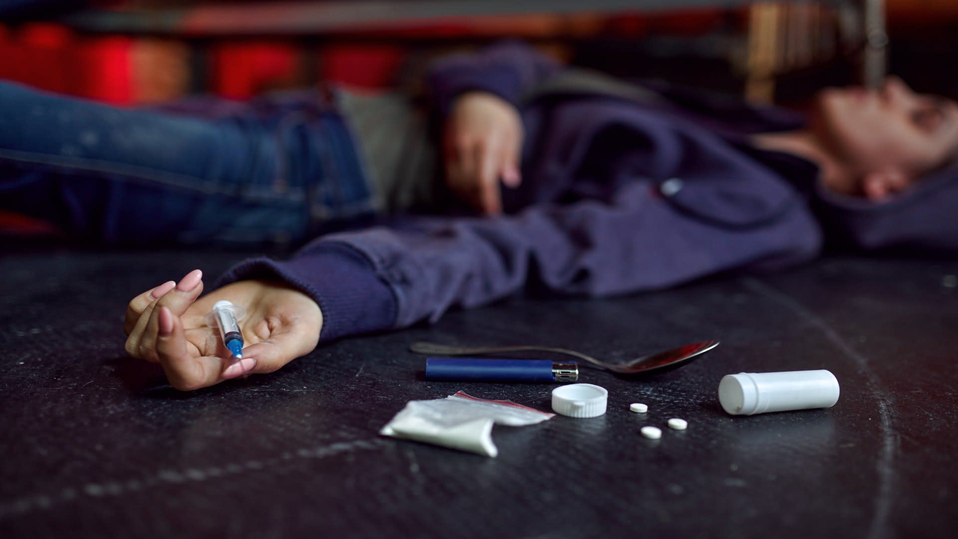 Opioid overdose aftermath with a young woman lying unconscious on the floor, a syringe and pills visible beside her.