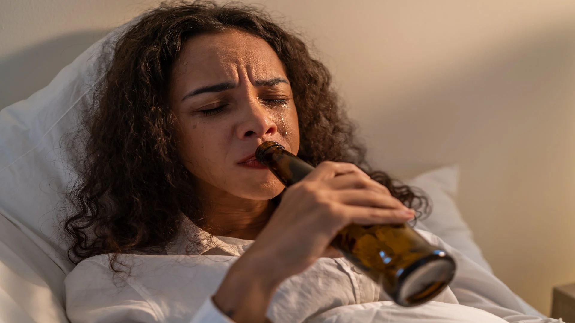 Alcohol intoxication symptoms shown in a distressed woman crying in bed while holding a beer bottle, conveying the emotional volatility often linked with intoxication.