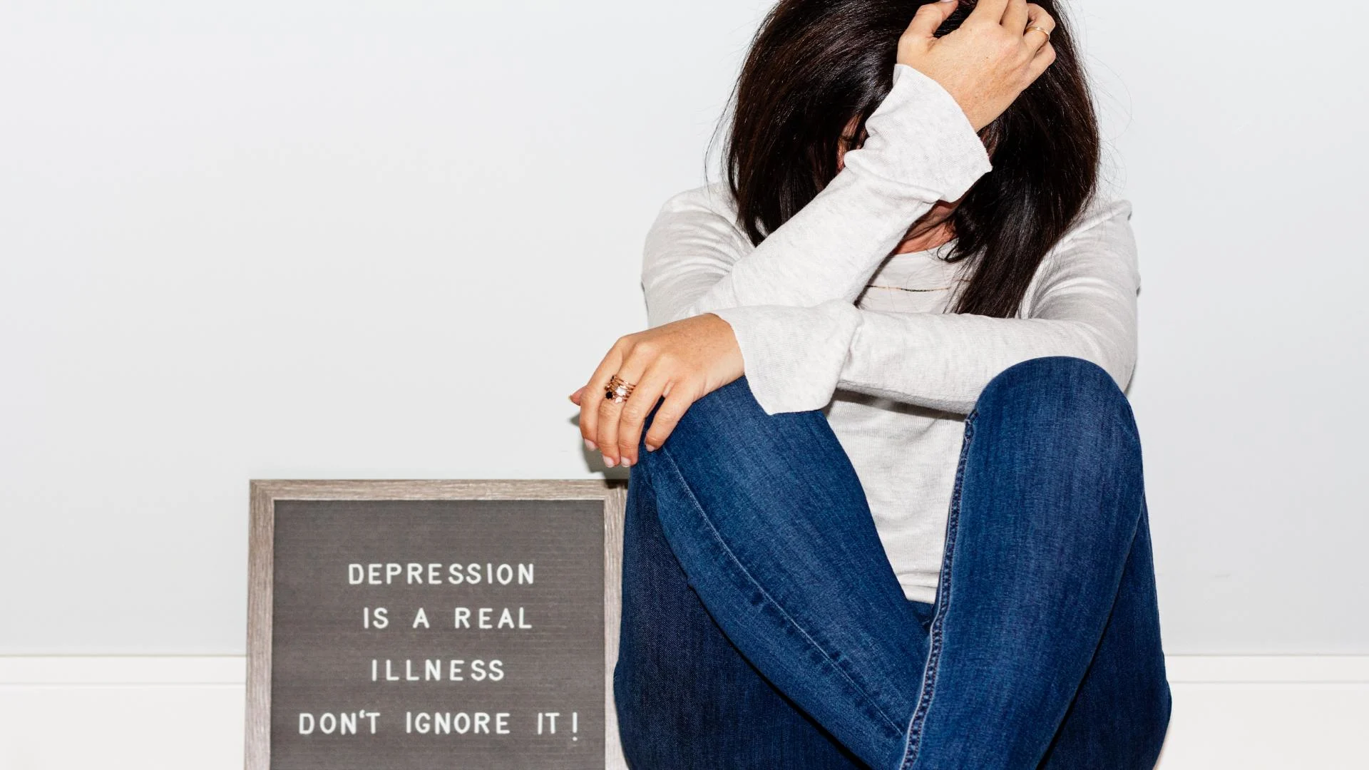 A woman sits on the floor with her head in her hand beside a sign that reads, “Depression is a real illness. Don’t ignore it!”—emphasizing the seriousness and need for awareness around mental health.