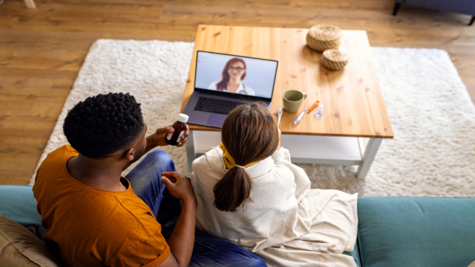 Couple sitting on a couch during a telehealth mental health session with a doctor visible on their laptop screen, illustrating remote care from home.
