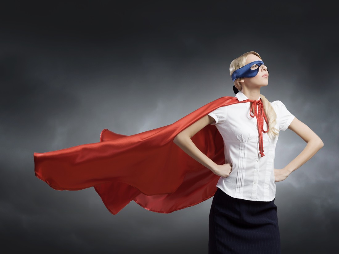 Woman wearing cape and mask and office clothes stands in superhero pose.