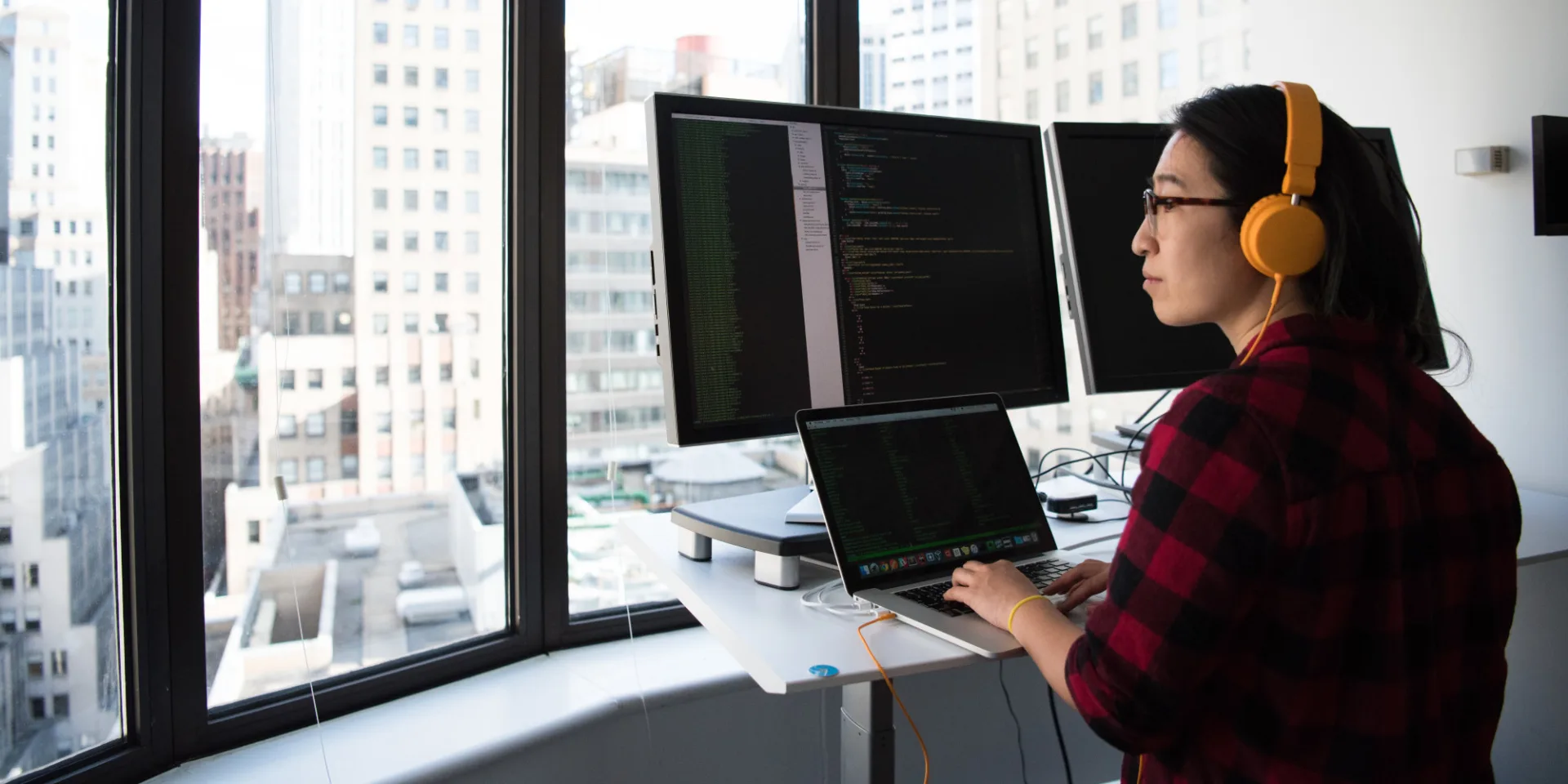 Woman coding on computer in front of large window with view of the city.