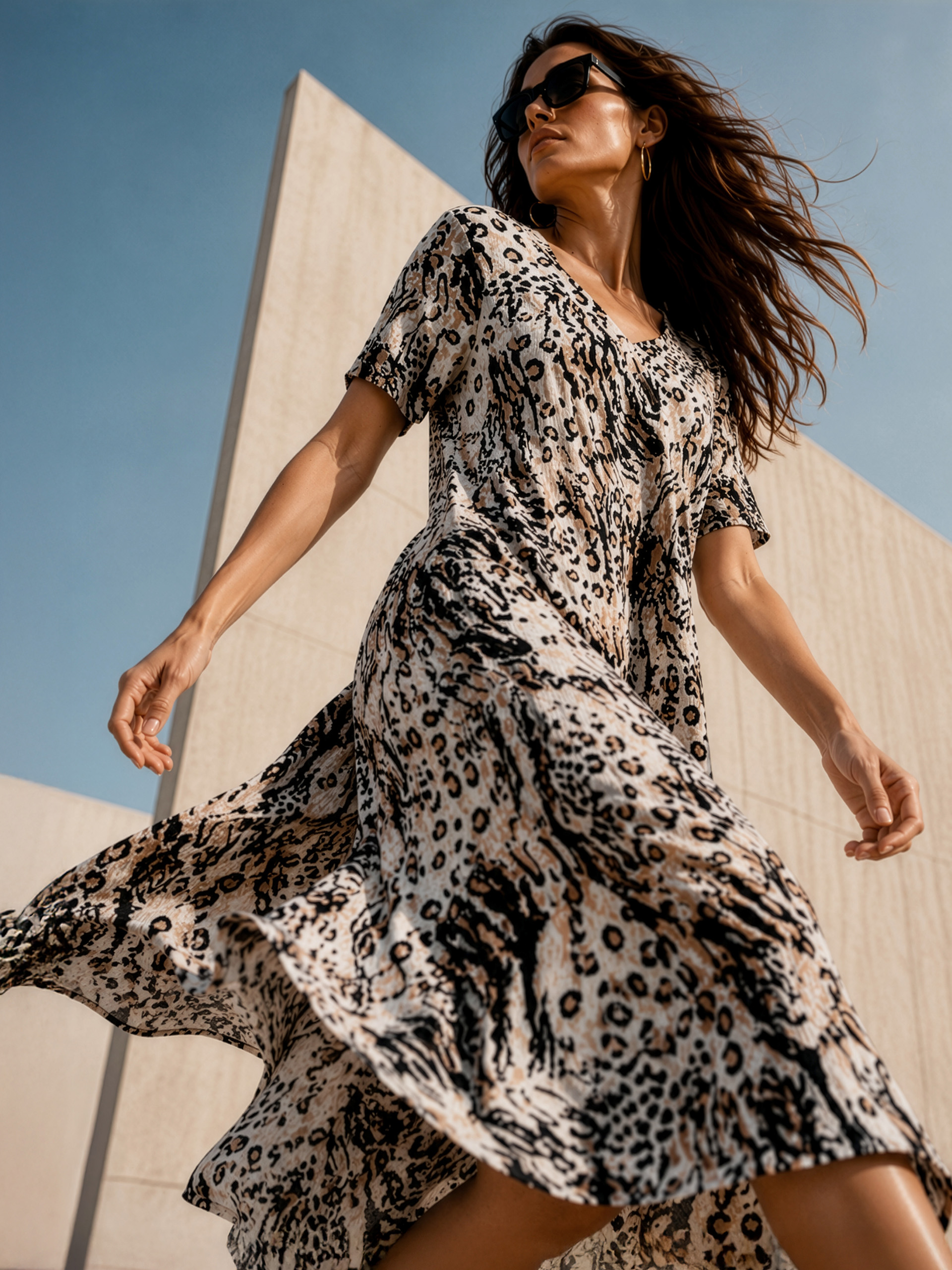 Ein Model trägt ein fließendes, kurzärmeliges Kleid mit Leopardenmuster und Sonnenbrille, posiert vor einer hellen Wand bei blauem Himmel.