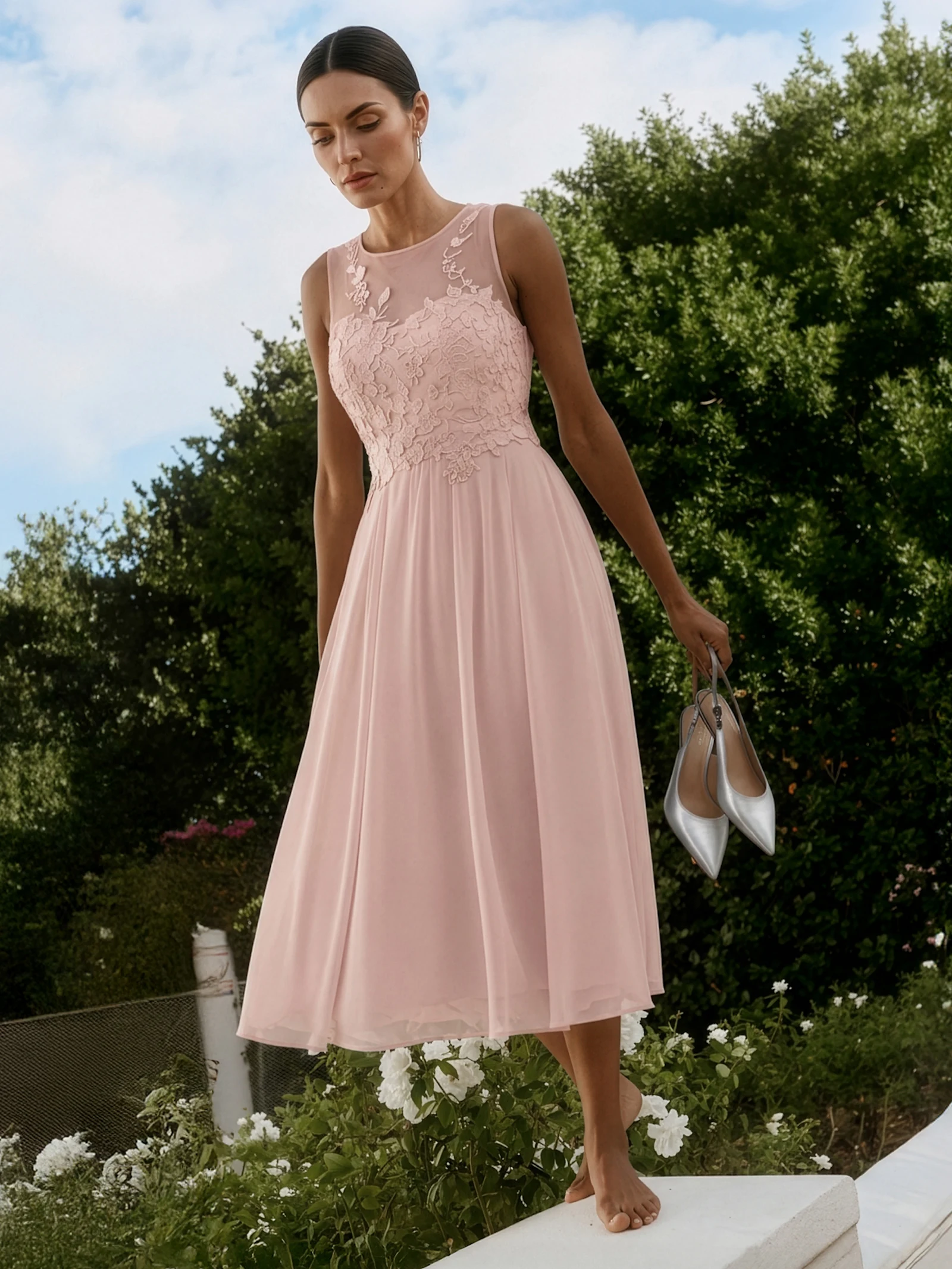 Un mannequin porte une robe rose sans manches avec un haut en dentelle et une jupe fluide, se tient pieds nus dans le jardin et tient des chaussures argentées à la main.