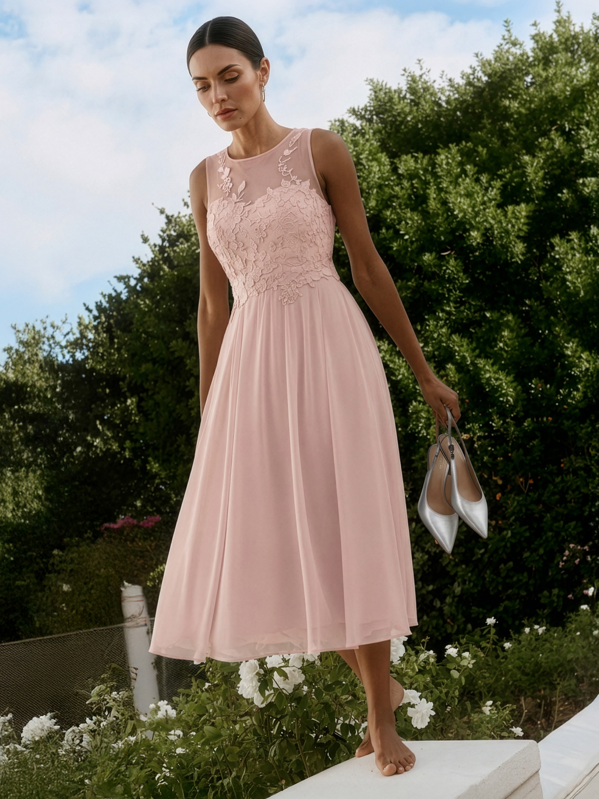 Een model draagt een mouwloze, roze jurk met een kanten bovenstuk en een zwierige rok, staat blootsvoets in de tuin en houdt zilveren schoenen in de hand.