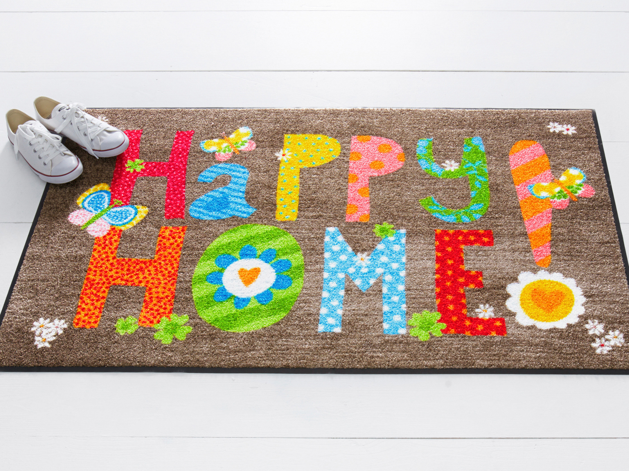 Eine Fußmatte zeigt den bunten Schriftzug "Happy Home!" in lebhaften Farben und Mustern. Neben der Matte sind Teile von weißen Schuhen zu sehen.