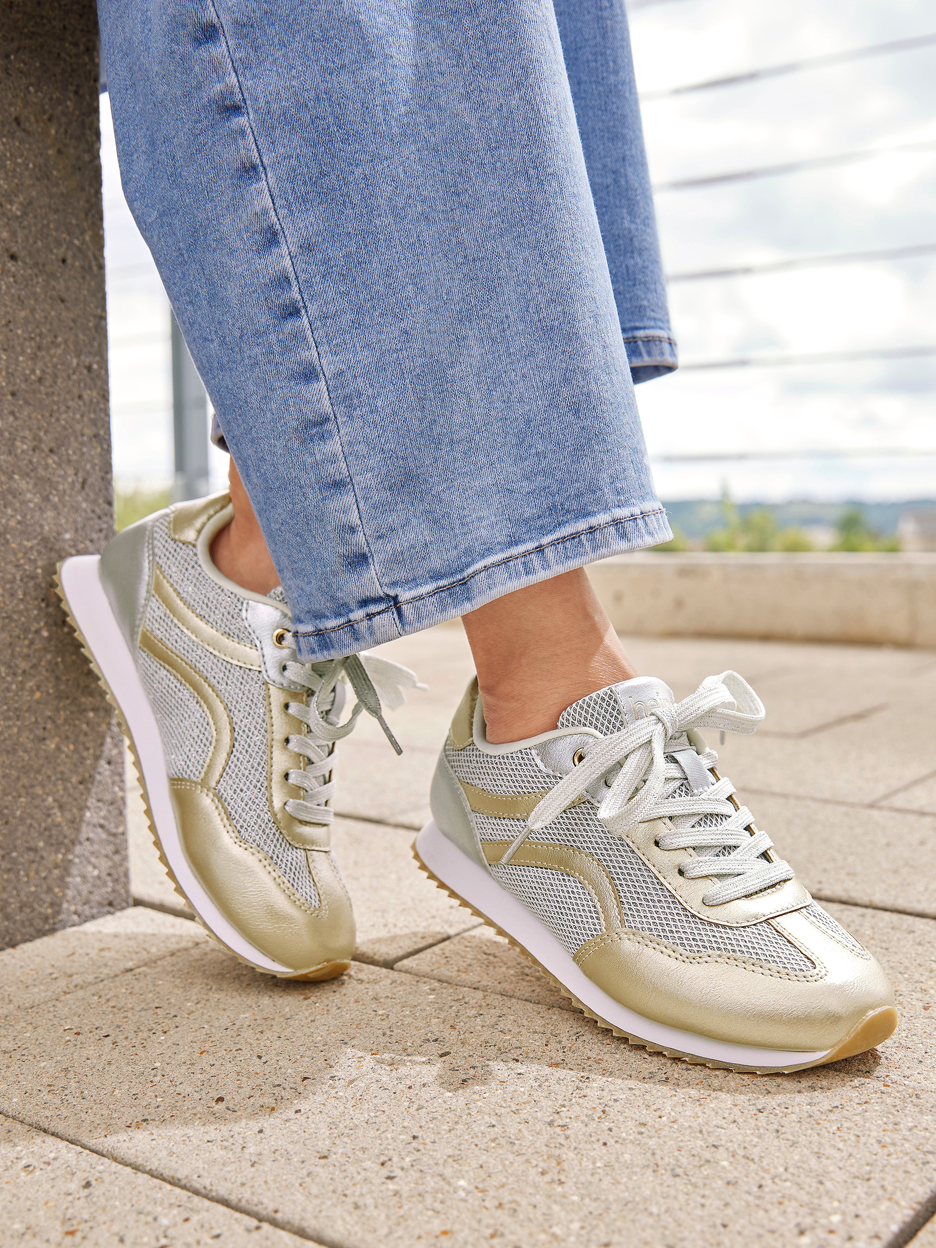 Nahaufnahme von silber-goldenen Sneakern mit weißen Schnürsenkeln, getragen zu blauen Jeans, auf einem gepflasterten Boden.