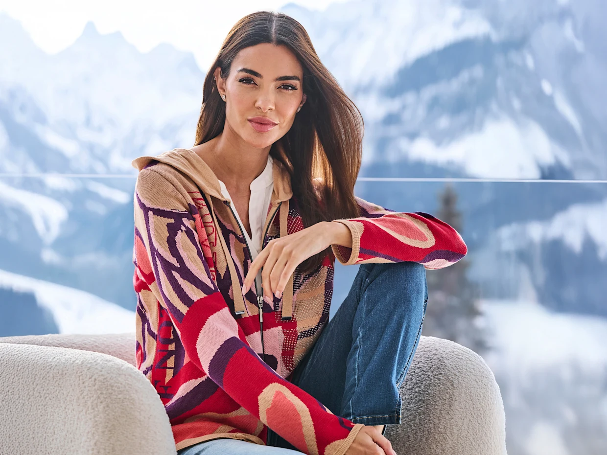 Een model zit ontspannen op een lichte stoel, draagt een kleurrijk gebreid vest en een blauwe spijkerbroek, met besneeuwde bergen op de achtergrond. Een model zit ontspannen op een lichte stoel, draagt een kleurrijk gebreid vest en een blauwe spijkerbroek, met besneeuwde bergen op de achtergrond.