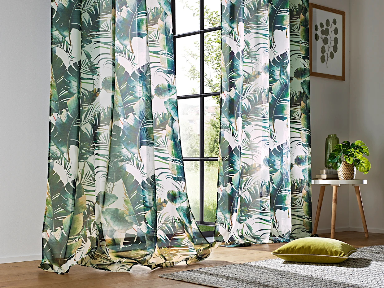 Grosse Fenster sind mit Gardinen in einem tropischen Blattmuster dekoriert. Ein grünes Kissen liegt auf einem Teppich, und eine Pflanze steht auf einem kleinen Tisch.