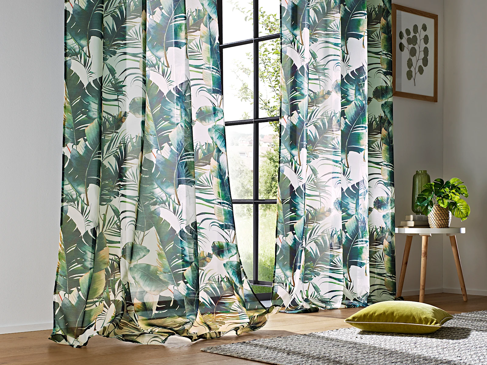 De grandes fenêtres sont décorées de rideaux dans un motif de feuilles tropicales. Un coussin vert est posé sur un tapis et une plante est posée sur une petite table.