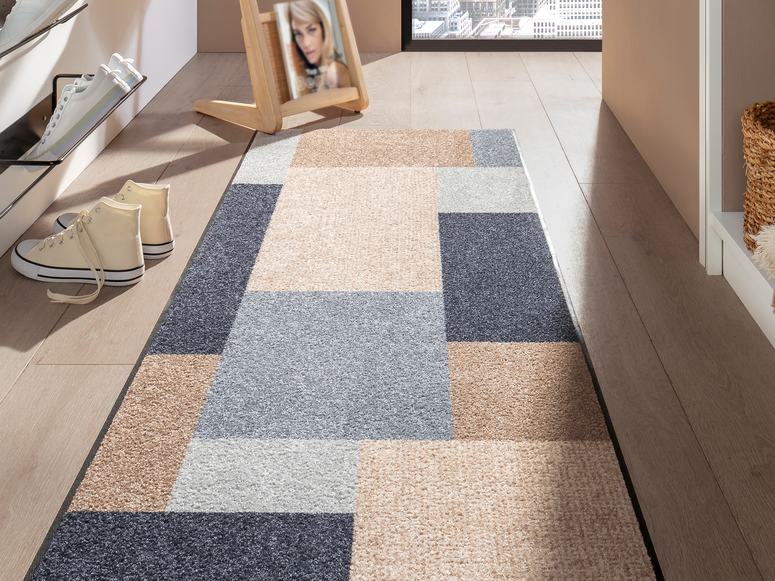 Un tapis à motif géométrique en tons beige, gris et bleu est posé sur un sol en bois clair, à côté deux paires de chaussures et un miroir.