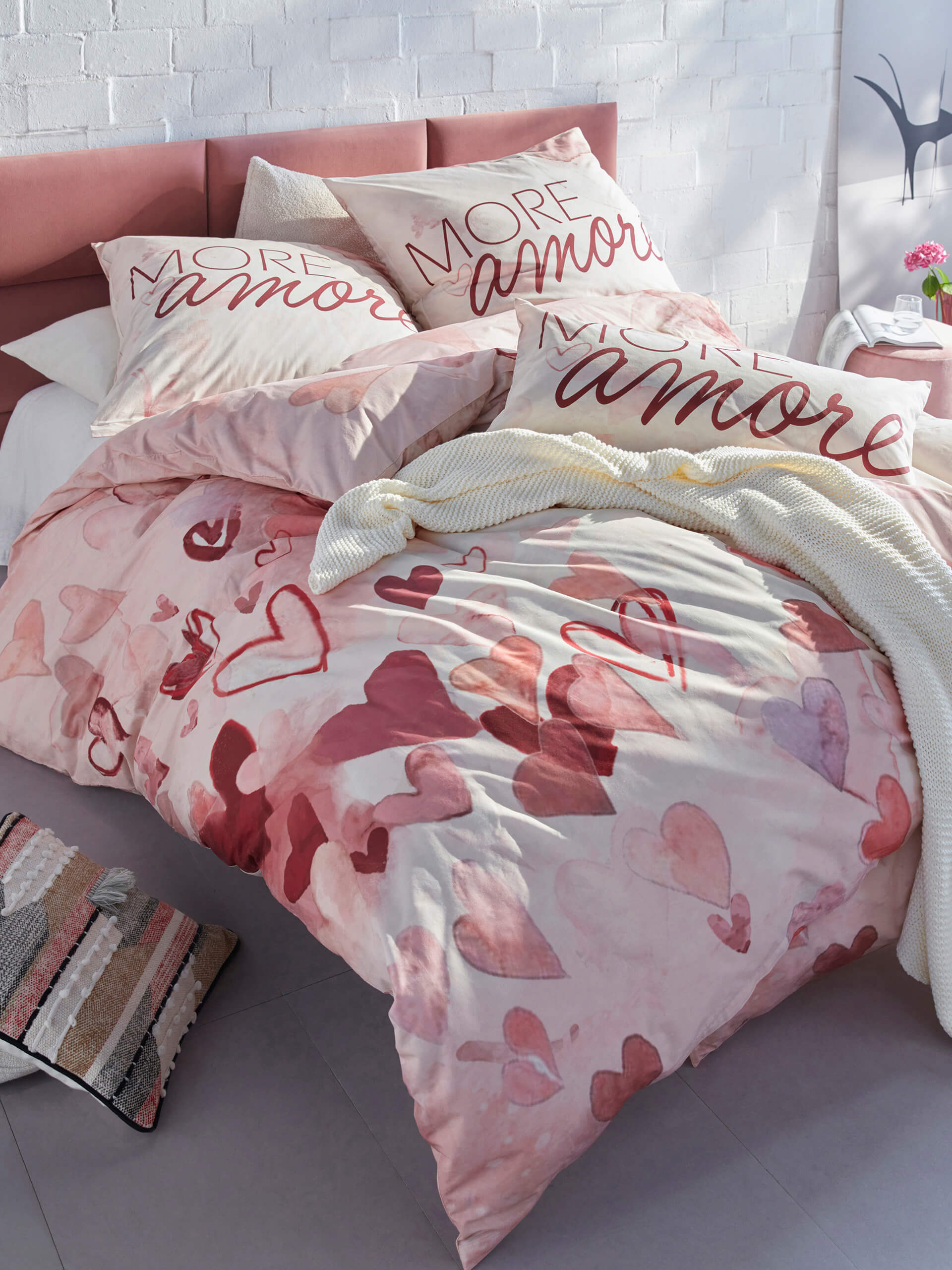 Un lit avec une literie rose à motif de cœurs et des coussins avec l'inscription 'More amor'. À côté, deux tapis roses et beiges avec un motif floral en relief.