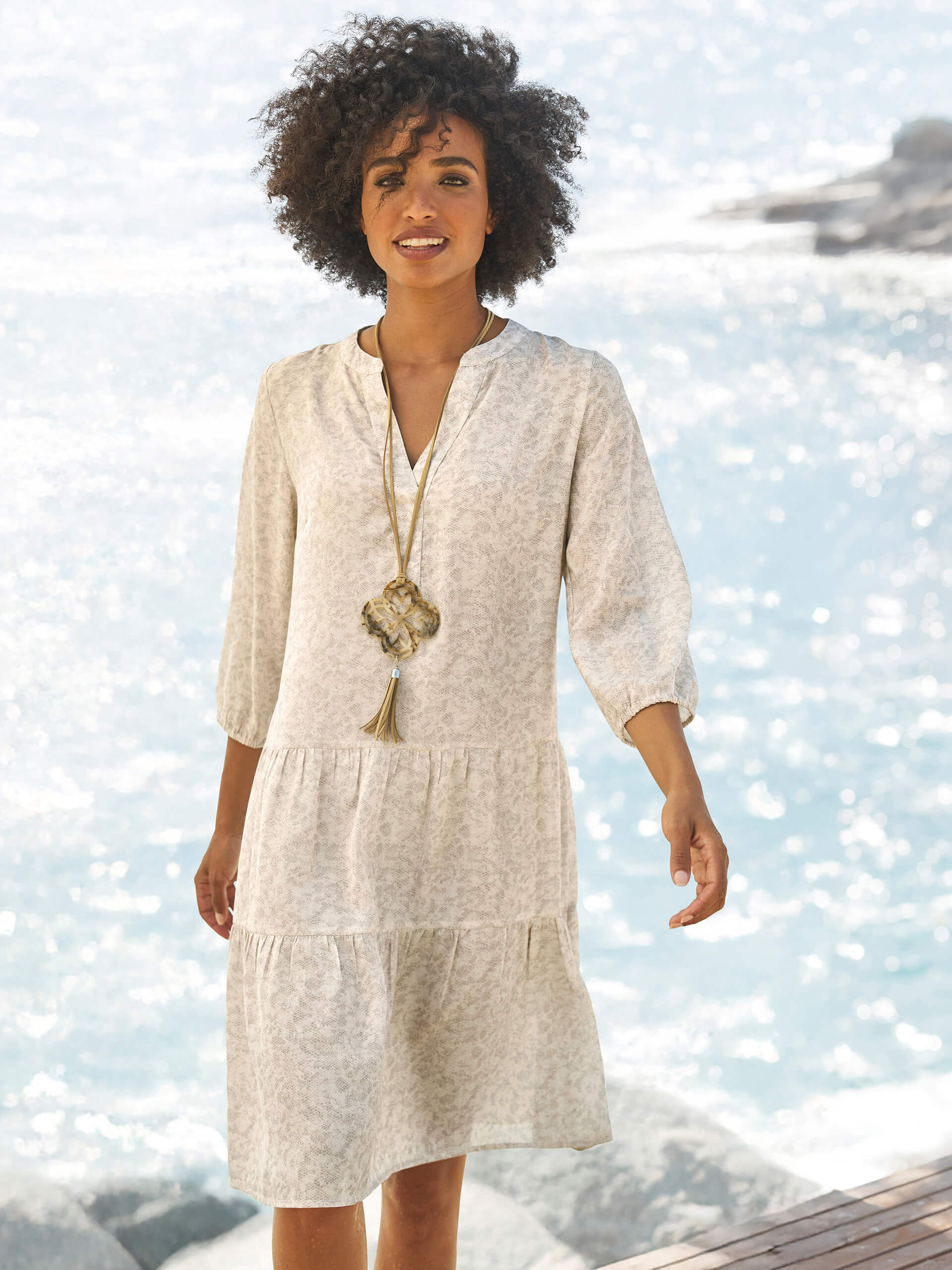 Un modèle se tient près de l'eau et porte une robe claire, longueur genou, à manches 3/4 et motif fin, ainsi qu'un long collier.