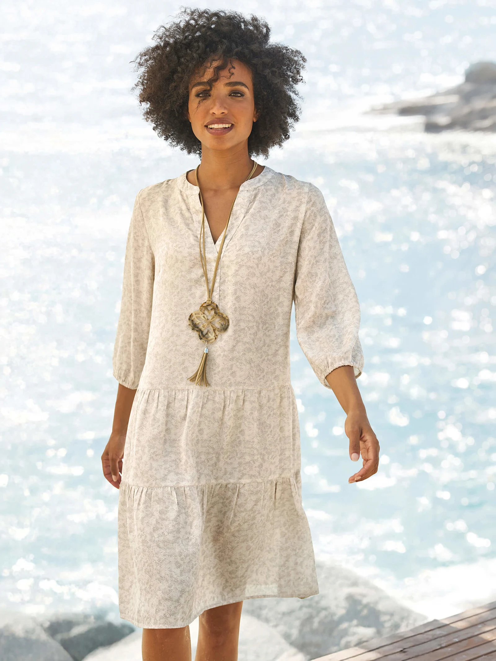 Ein Model steht am Wasser und trägt ein helles, knielanges Kleid mit 3/4-Ärmeln und feinem Muster sowie eine lange Kette.