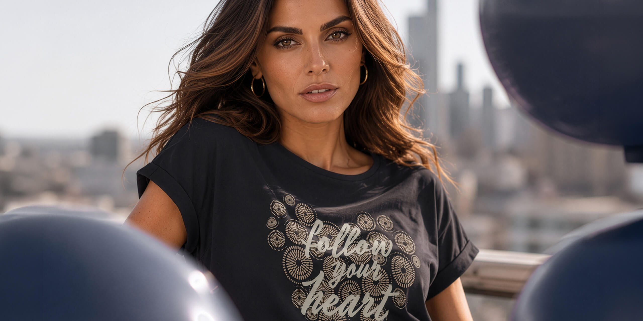 Ein Model trägt ein schwarzes T-Shirt mit goldfarbenem Herz und Schriftzug 'follow your heart', im Hintergrund Stadt und große schwarze Luftballons.