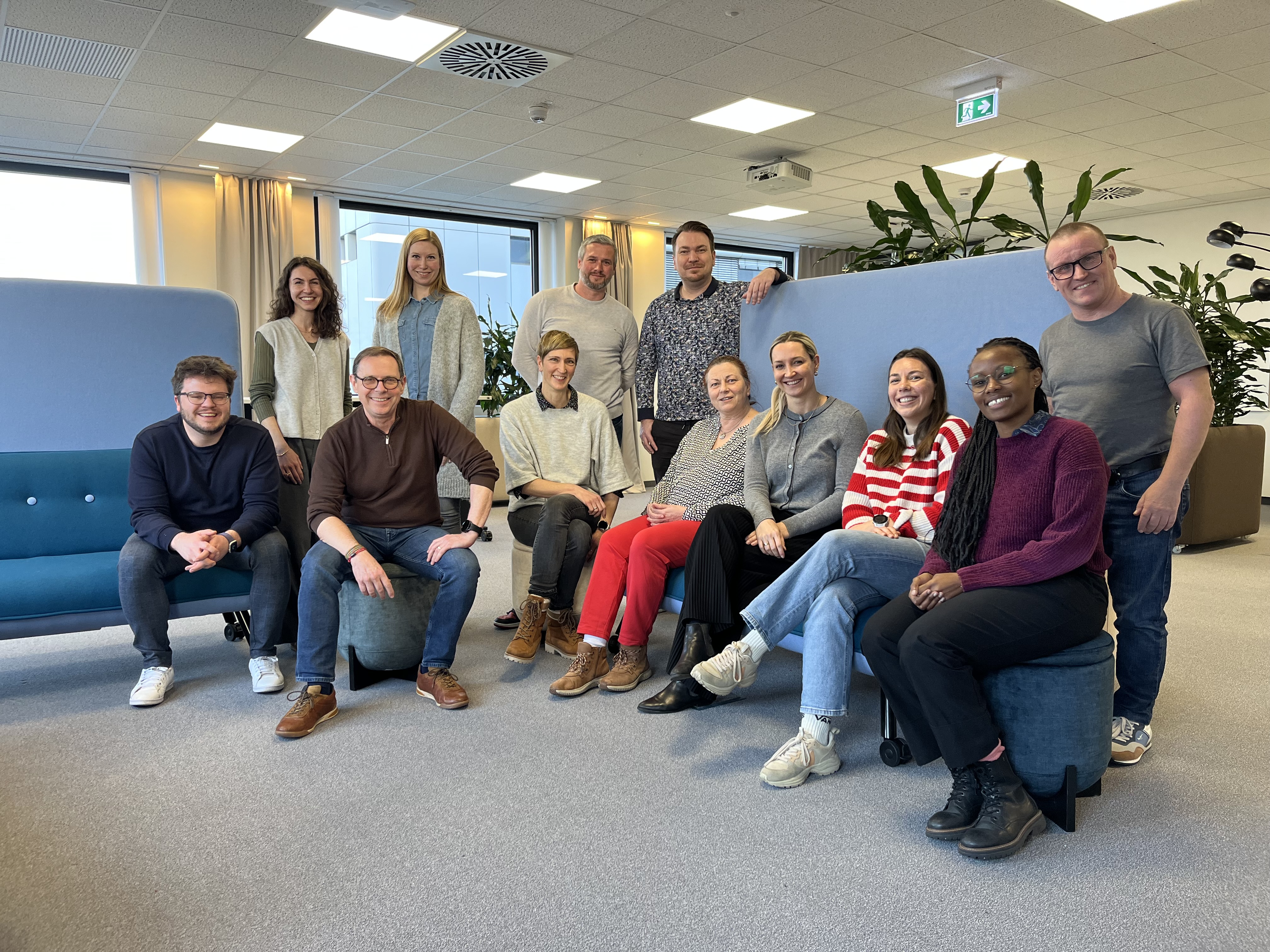 Een groep mensen zit en staat in een moderne kantoorruimte met blauwe meubels en groene planten op de achtergrond.