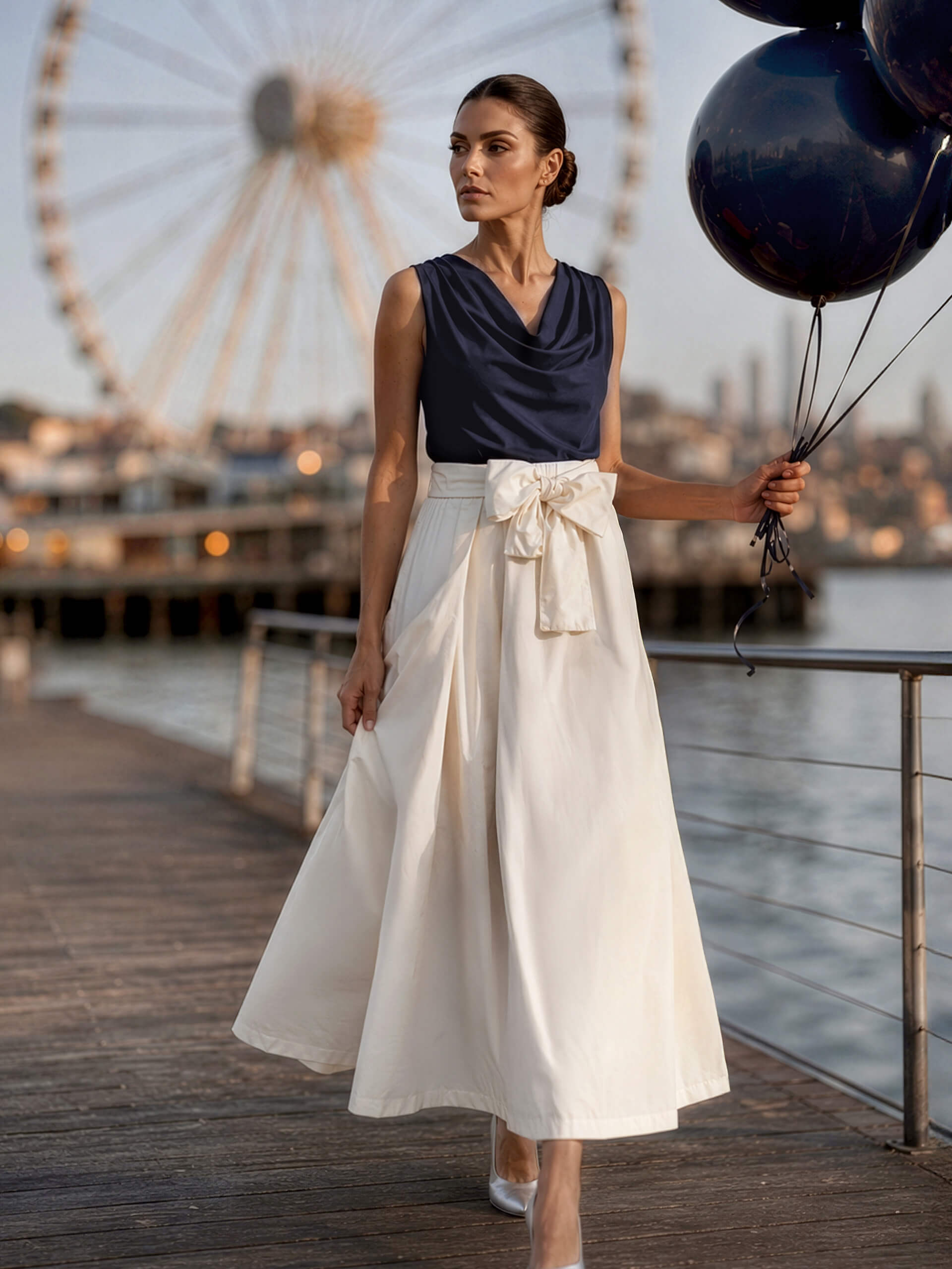 Un mannequin sur une jetée porte un haut bleu marine et une jupe blanche, tient des ballons noirs ; grande roue en arrière-plan.