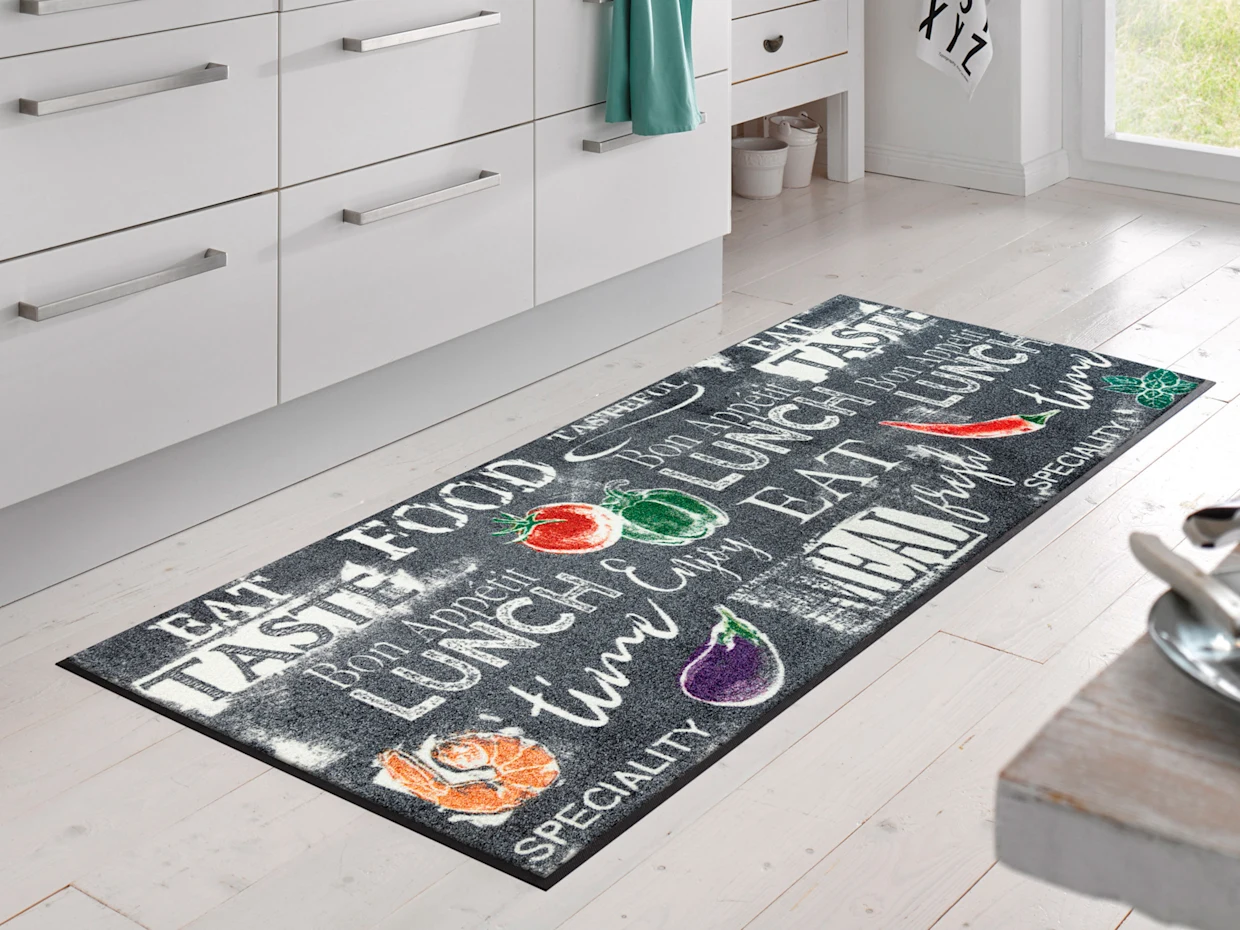 Een keuken met witte kastjes is bedekt met een lang tapijt met verschillende etenswaren en letters in felle kleuren. De lichte houten vloer en het licht dat door het raam naar binnen valt, geven de kamer een vriendelijke sfeer.