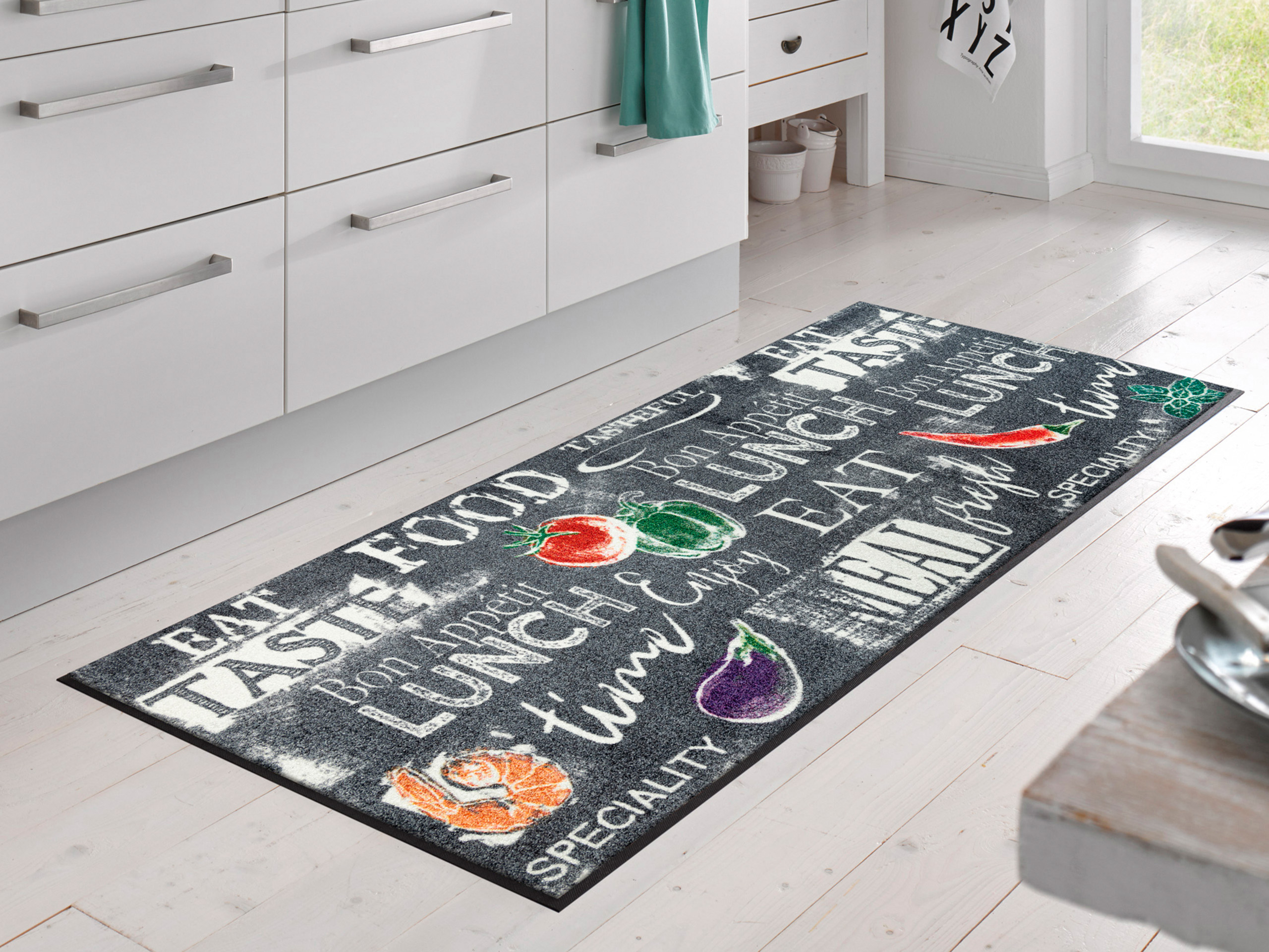 Een keuken met witte kastjes is bedekt met een lang tapijt met verschillende etenswaren en letters in felle kleuren. De lichte houten vloer en het licht dat door het raam naar binnen valt, geven de kamer een vriendelijke sfeer.