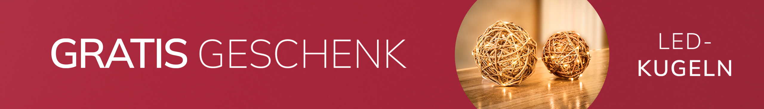 Eine Werbeanzeige mit bordeauxrotem Hintergrund, mit dem Text &#x27;GRATIS GESCHENK*&#x27;
Daneben sieht man zwei leuchtende Dekokugeln auf einem Holztisch.