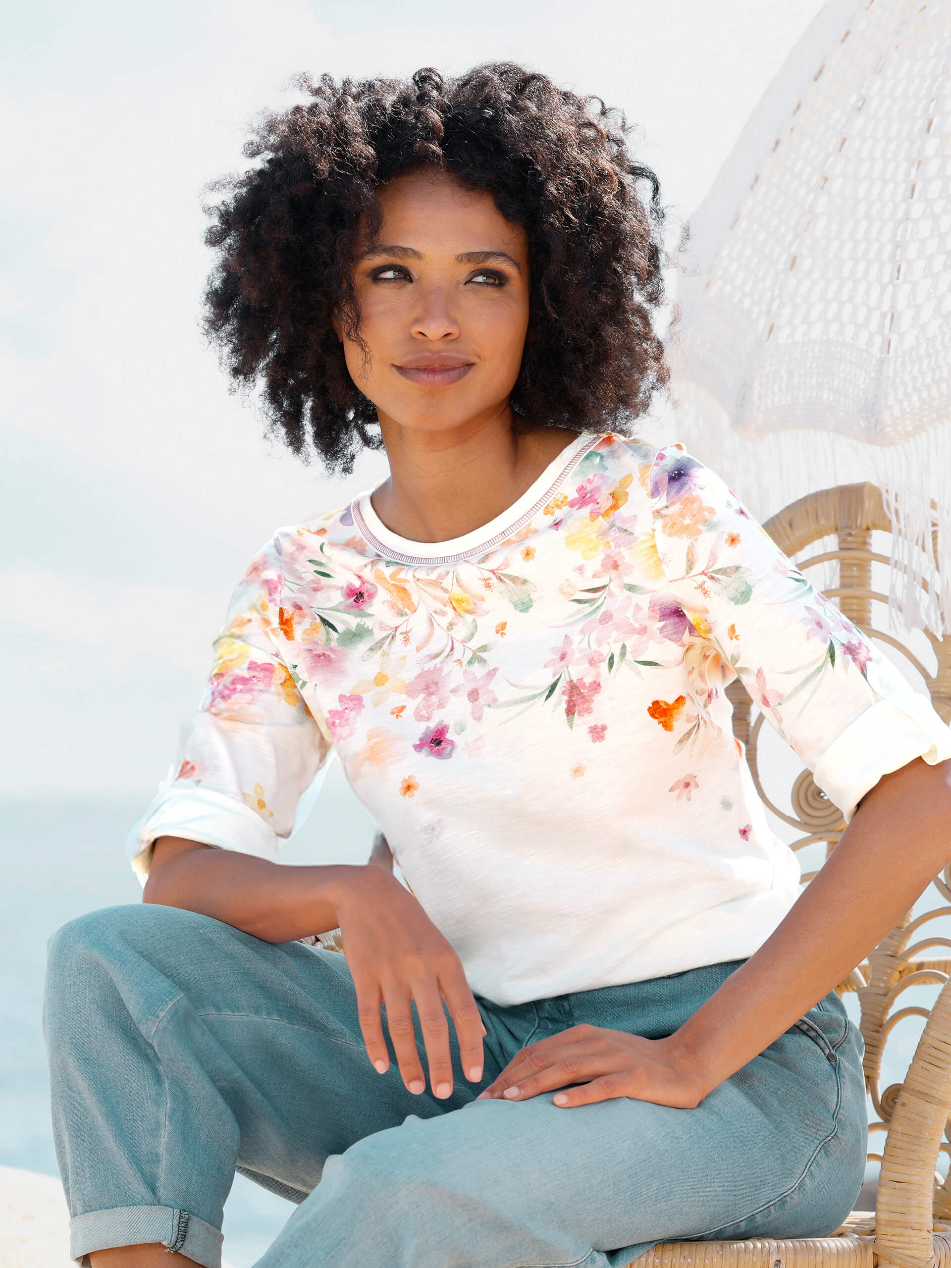 Ein Model sitzt auf einem Stuhl am Strand, trägt ein weißes Shirt mit buntem Blumenmuster und eine hellblaue Jeans.