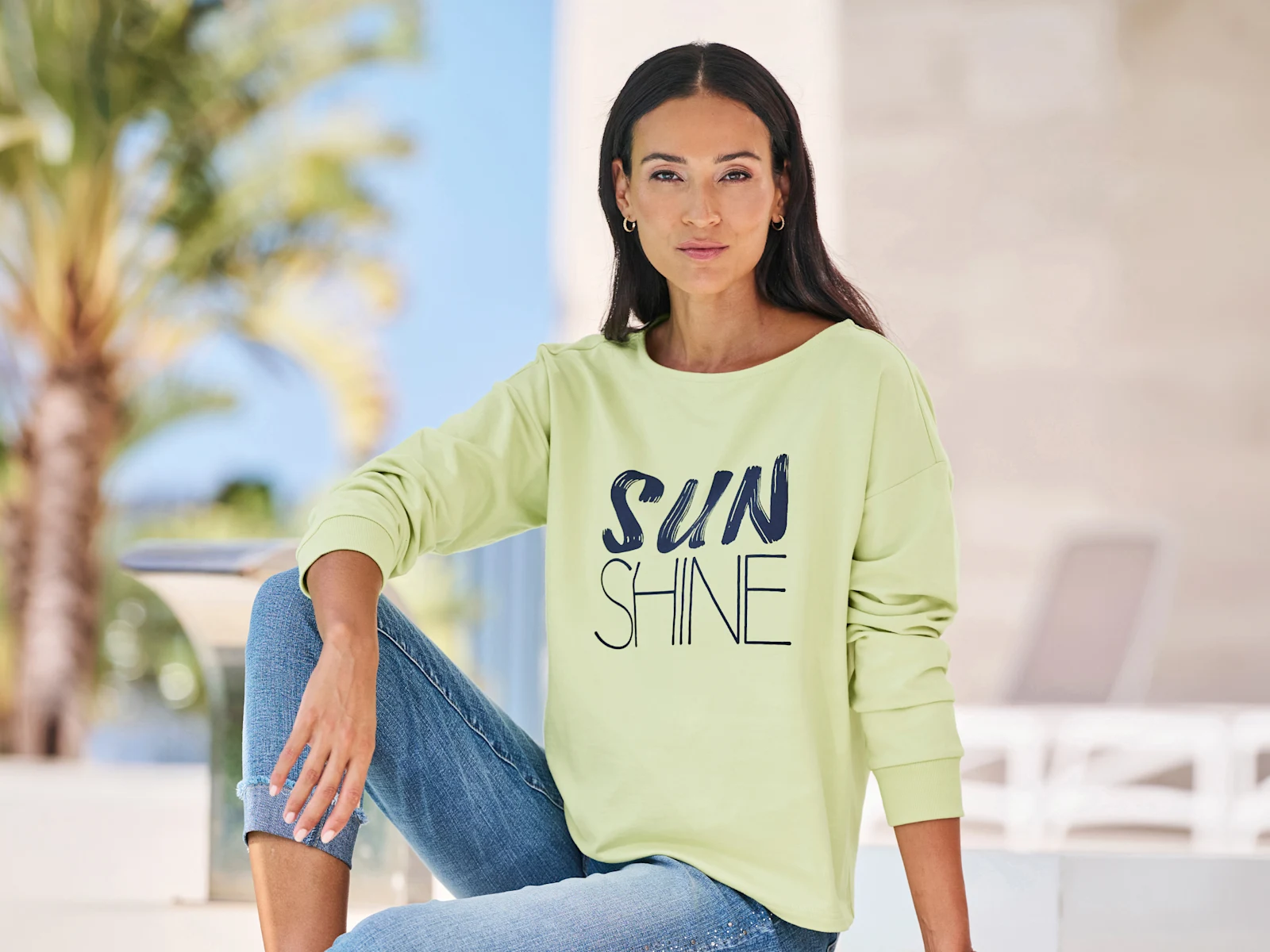 Un modèle est assis détendu, portant un sweat-shirt vert clair avec l'inscription 'SUNSHINE' et un jean bleu.