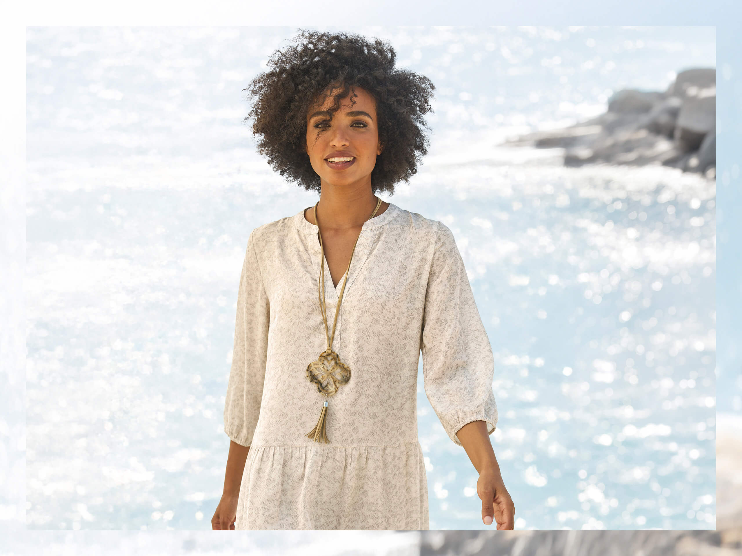 Un modèle se tient près de l'eau et porte une robe claire, longueur genou, à manches 3/4 et motif fin, ainsi qu'un long collier.