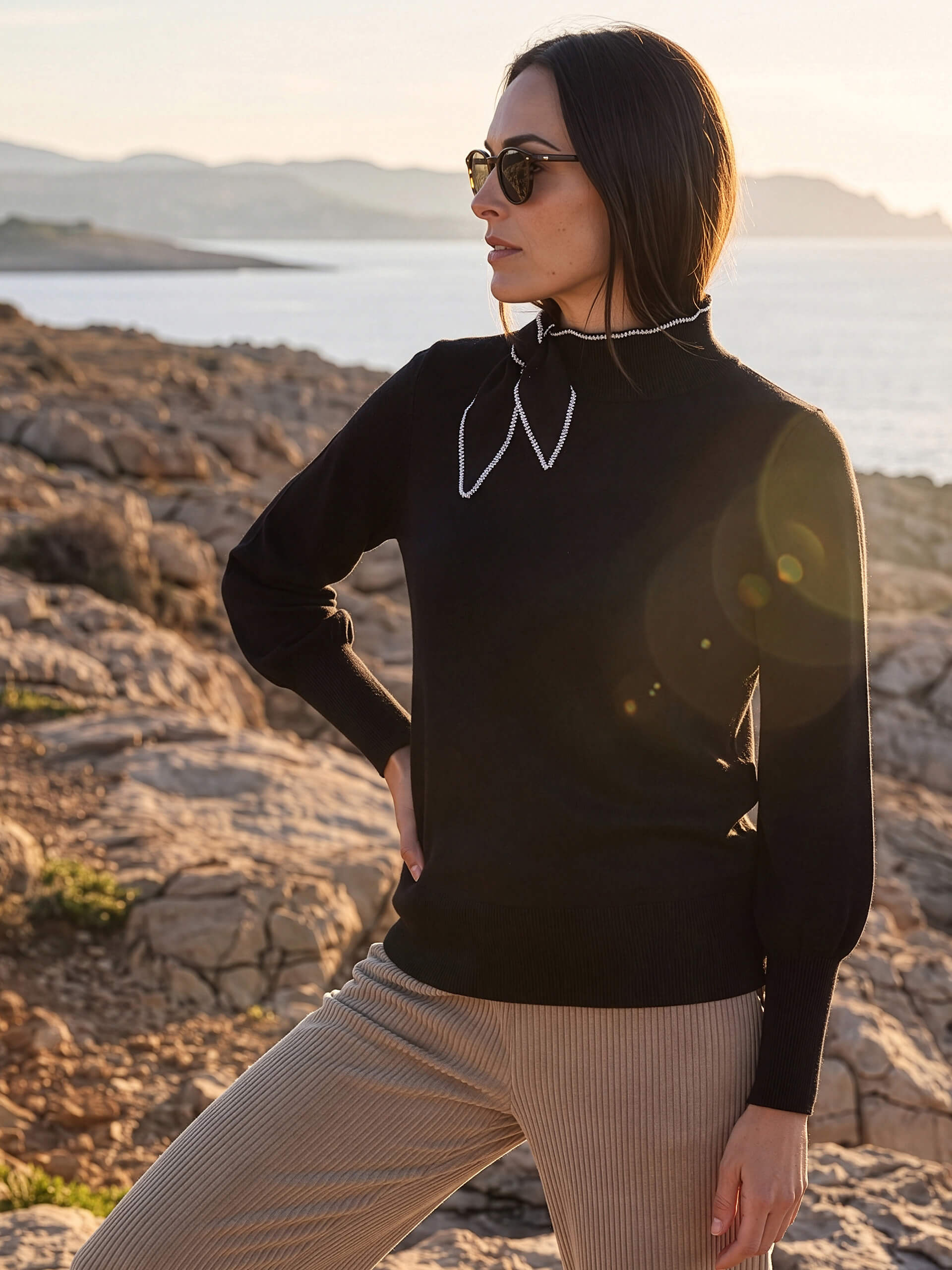 Ein Model in schwarzem Pullover mit Schleifendetail und beiger Hose steht mit Sonnenbrille in einer felsigen Küstenlandschaft.