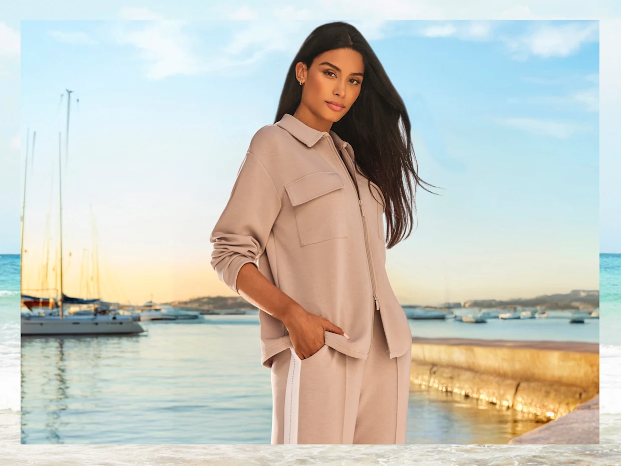 Ein Model steht am Hafen bei Sonnenuntergang und trägt einen beigefarbenen Hosenanzug mit weißen Sneakern.