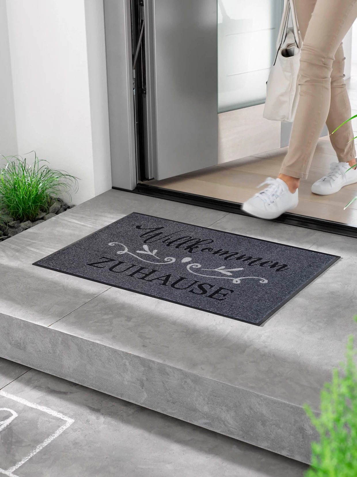 Ein Model betritt ein Haus und steht auf einer grauen Fußmatte mit der Aufschrift "Willkommen Zuhause". Die Eingangstür ist offen und der Boden im Innenbereich ist aus Holz.