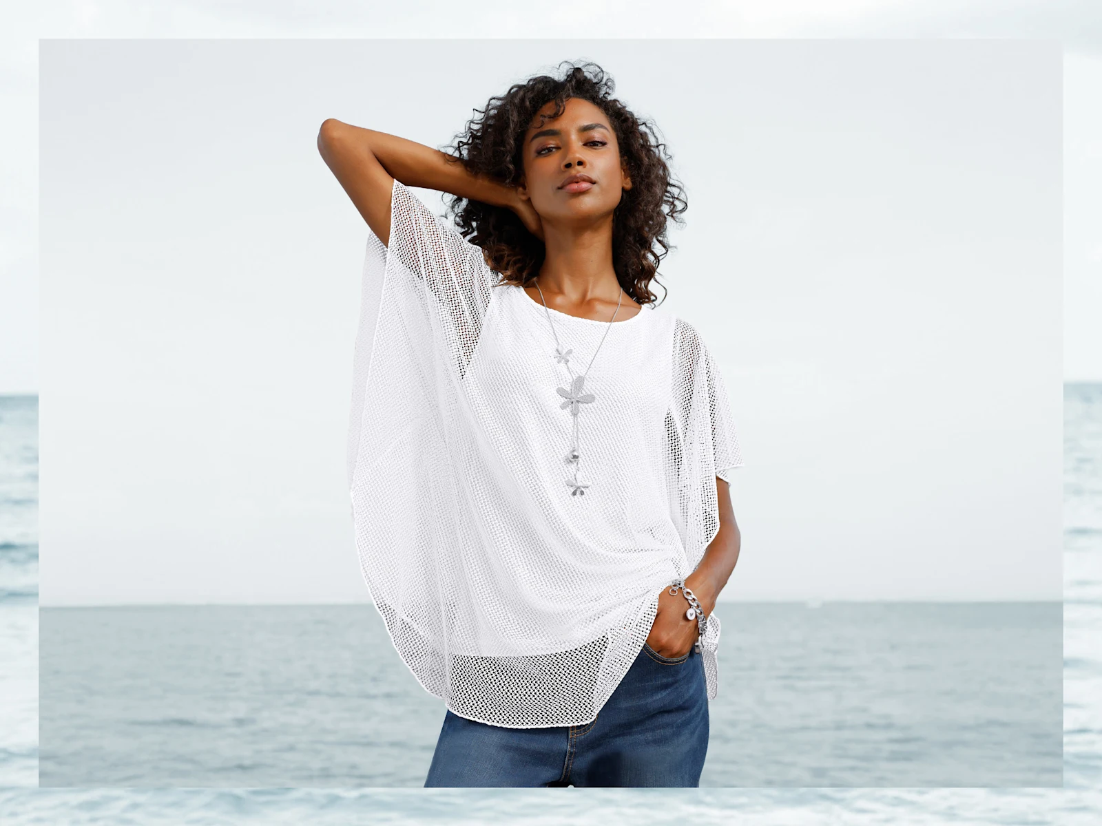 Een model draagt een wit, losvallend nettop met korte mouwen en sieraden, met de zee op de achtergrond.
