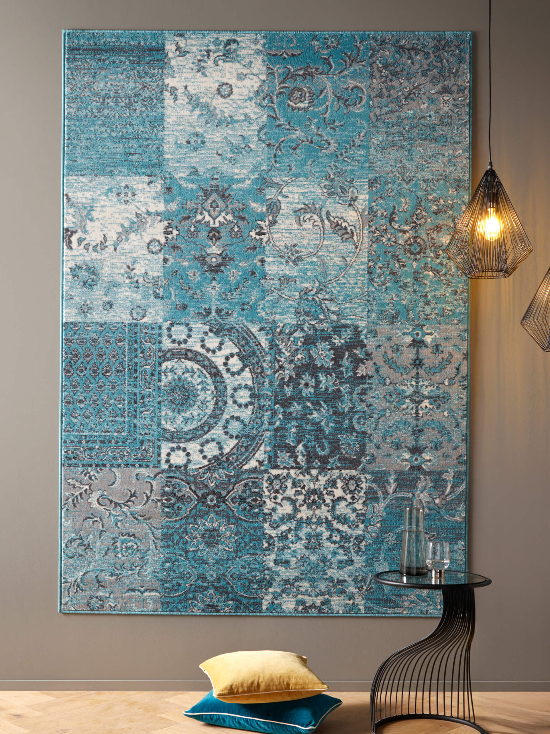 Wandkleed met blauw-grijs patchworkpatroon, daarnaast twee hangende draadlampen, kleine ronde tafel met glazen fles en twee kussens op de vloer.
