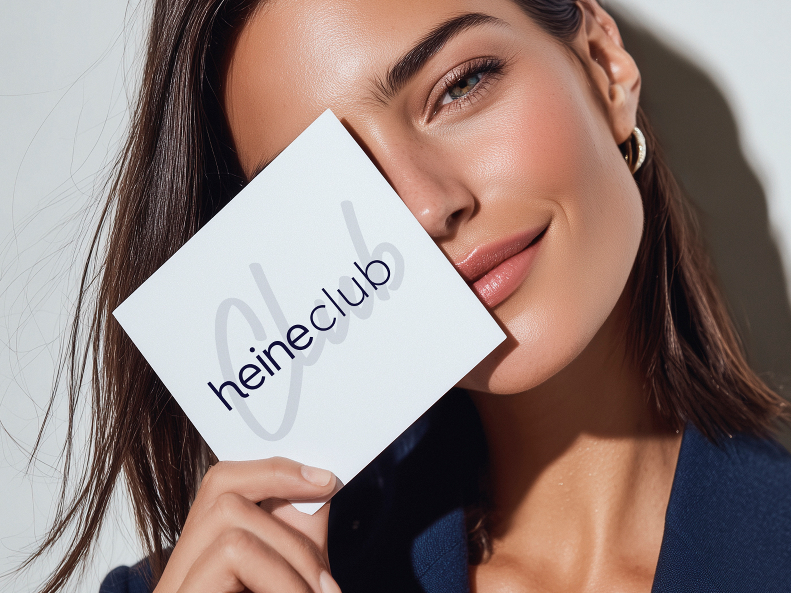 Ein Model hält eine Karte mit dem Text 'heineClub' vor das Gesicht, trägt dunkle Kleidung und goldene Ohrringe.