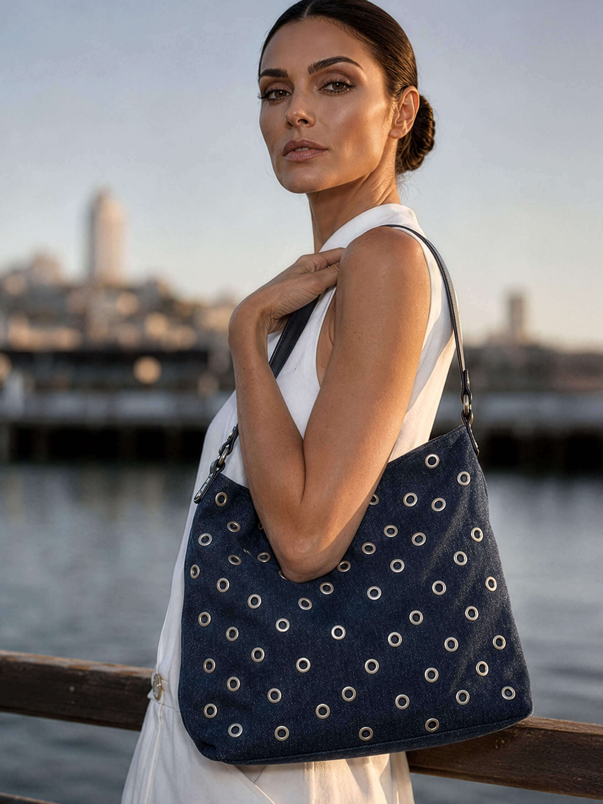 Ein Model am Wasser trägt ein weißes Kleid und eine dunkelblaue Schultertasche mit Nietenmuster.