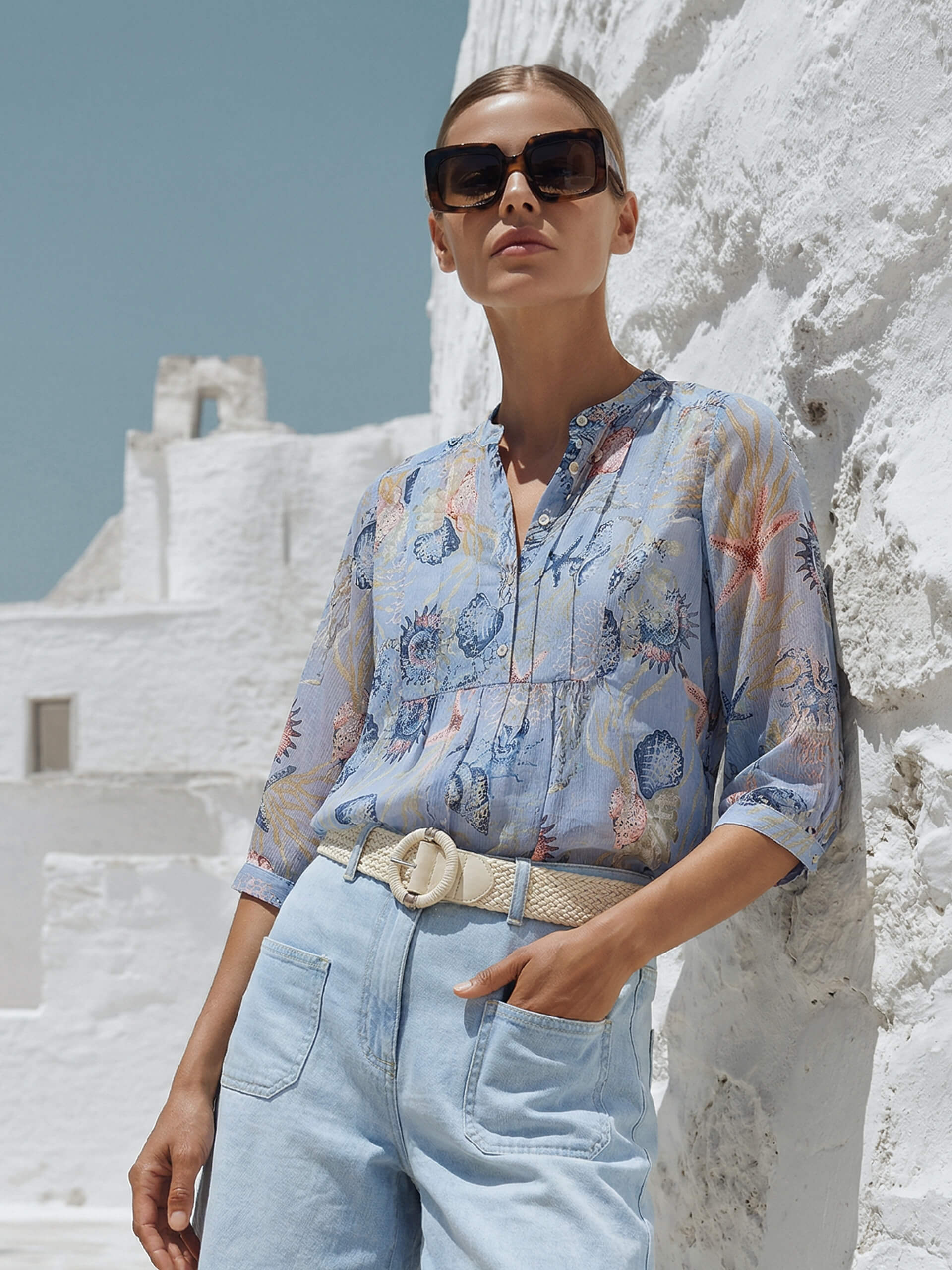 Un modèle appuyé contre un mur blanc, porte un chemisier clair à motifs avec manches trois-quarts et un jean clair avec ceinture.