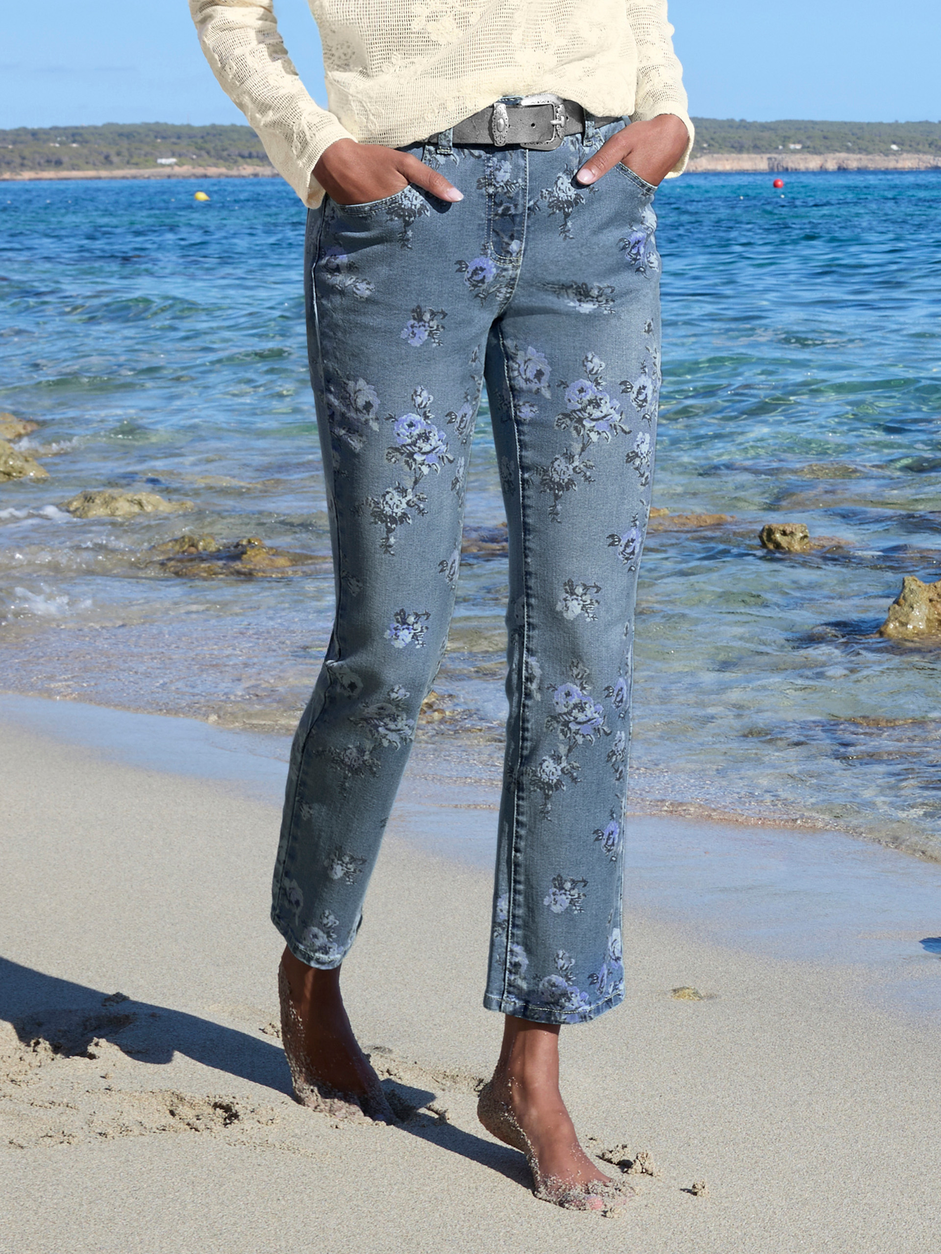 Un modèle aux cheveux bouclés porte un pull structuré crème et un jean bleu à motif floral, elle se tient au bord de la mer.