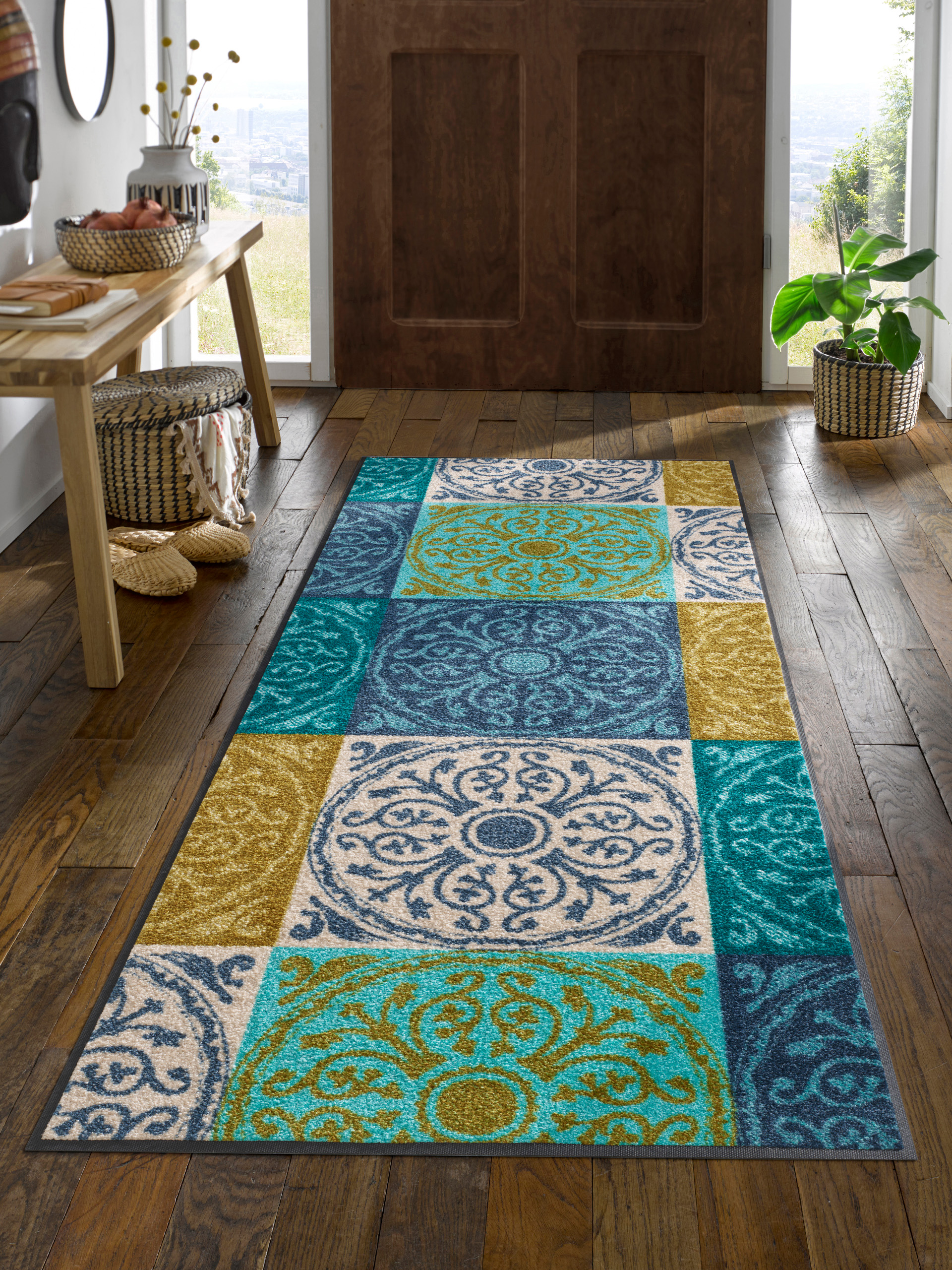 Un hall avec un sol en bois est décoré d'un long tapis rectangulaire dans différentes nuances de bleu, de vert et de jaune et de motifs décoratifs. Près d'une porte en bois se trouve une table avec des objets décoratifs et un panier avec une plante.