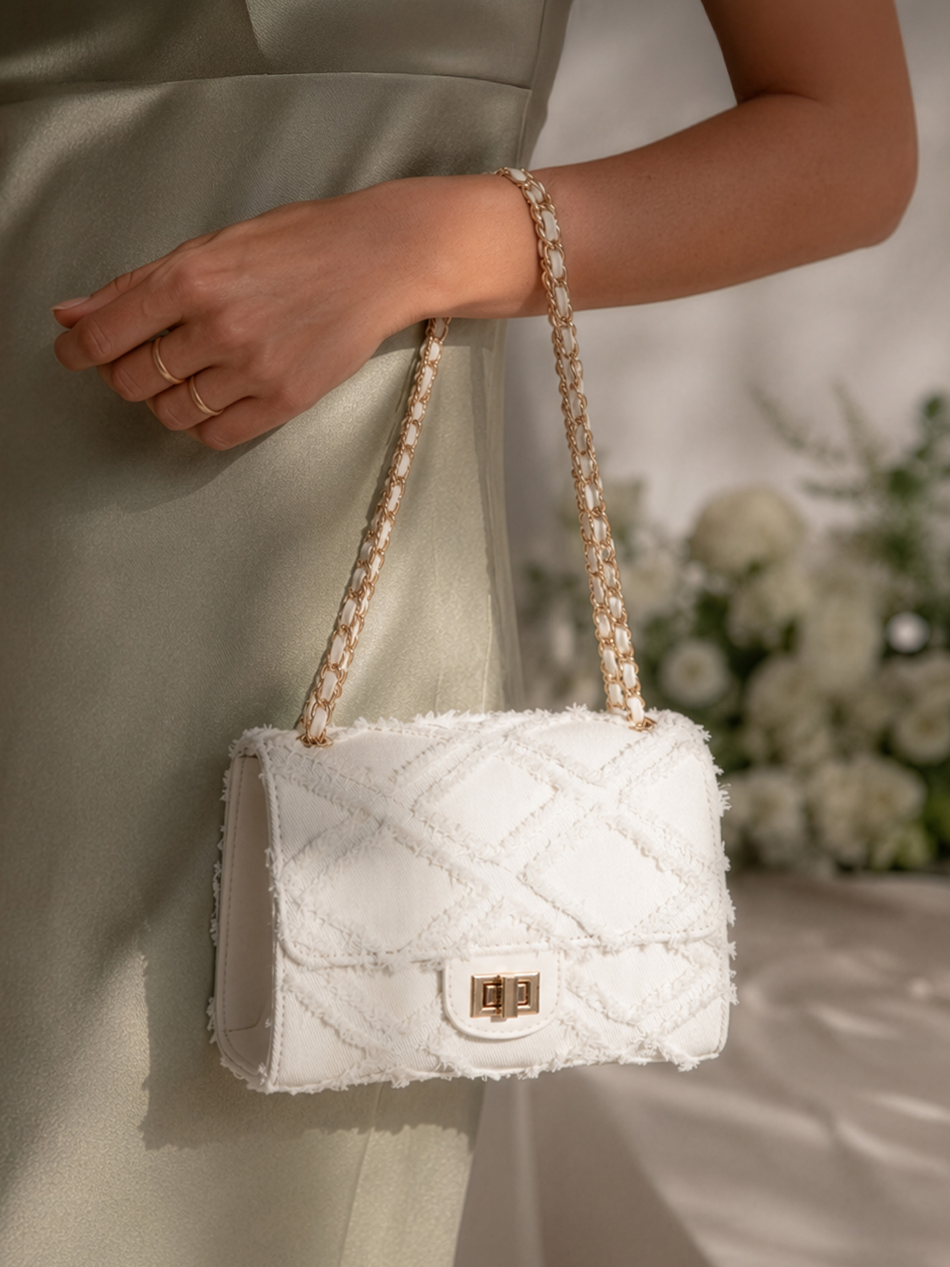 Une main ornée de bagues dorées tient un sac à main blanc texturé avec une chaîne dorée, le mannequin porte une robe vert clair, avec des fleurs blanches en arrière-plan.
