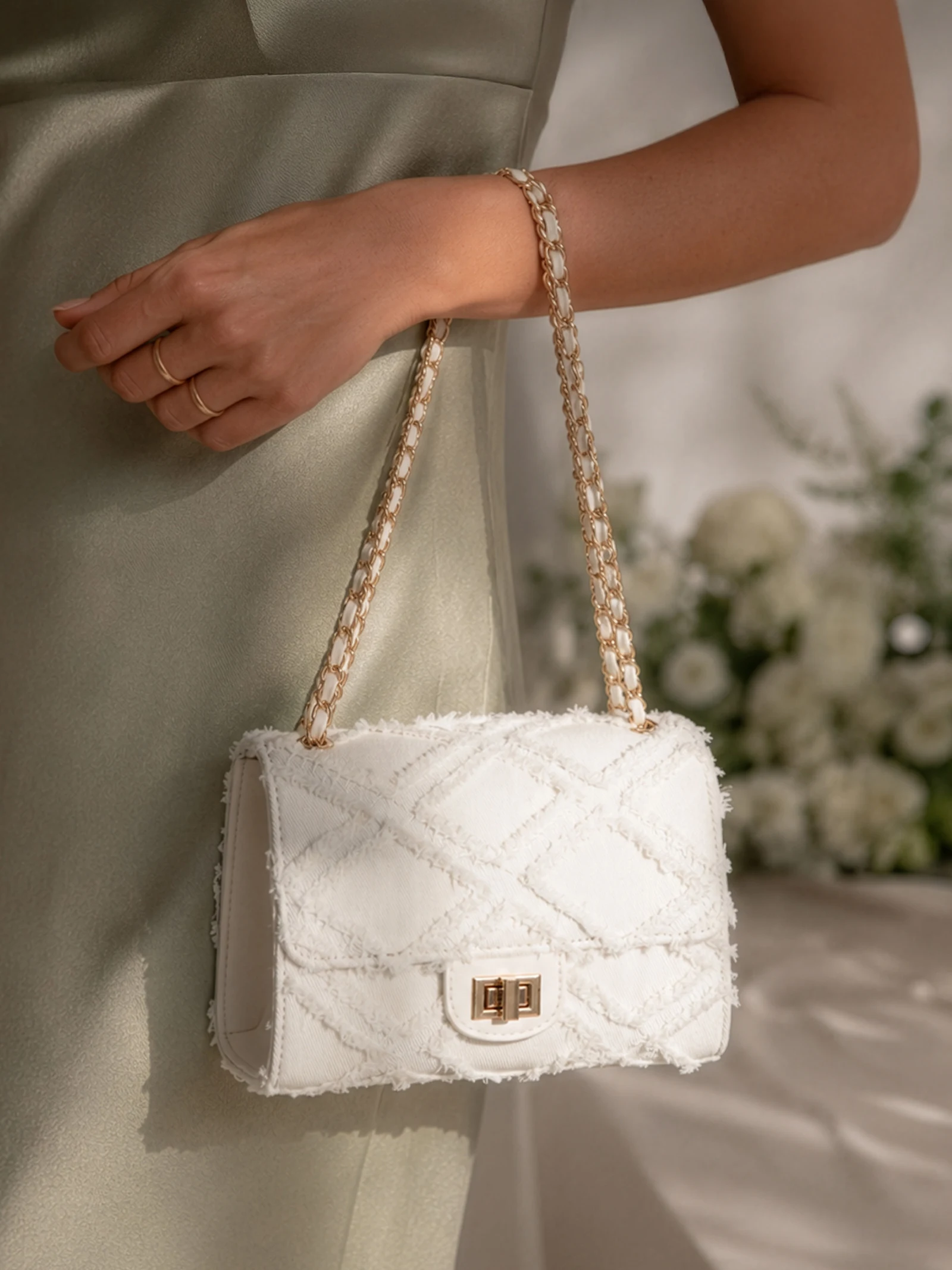 Une main ornée de bagues dorées tient un sac à main blanc texturé avec une chaîne dorée, le mannequin porte une robe vert clair, avec des fleurs blanches en arrière-plan.