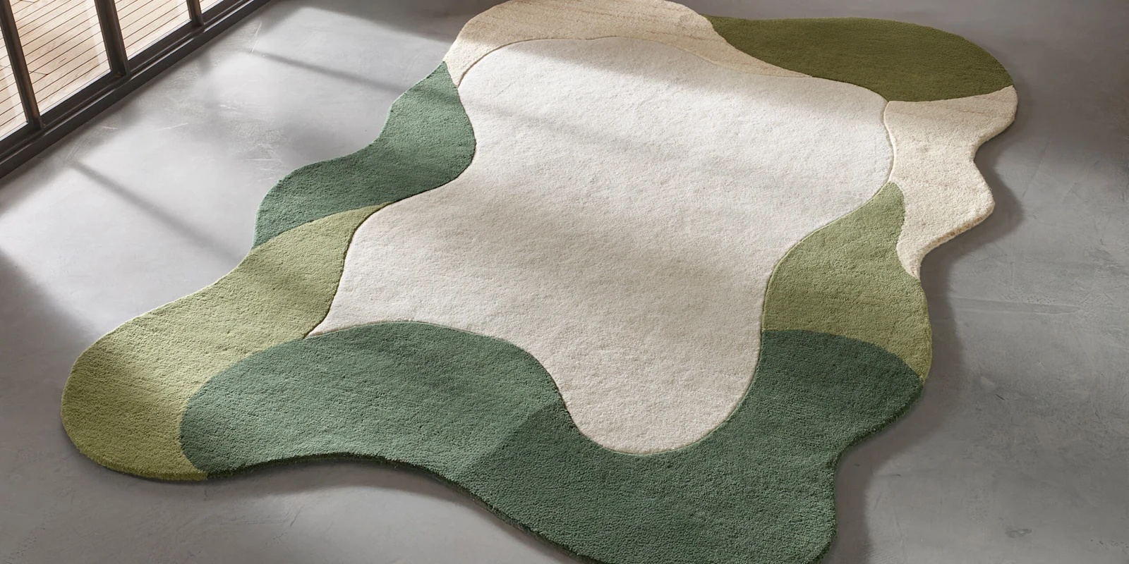 Moderne vloerkleed in organische vorm met vlakken in crème, lichtgroen en donkergroen op een grijze vloer.